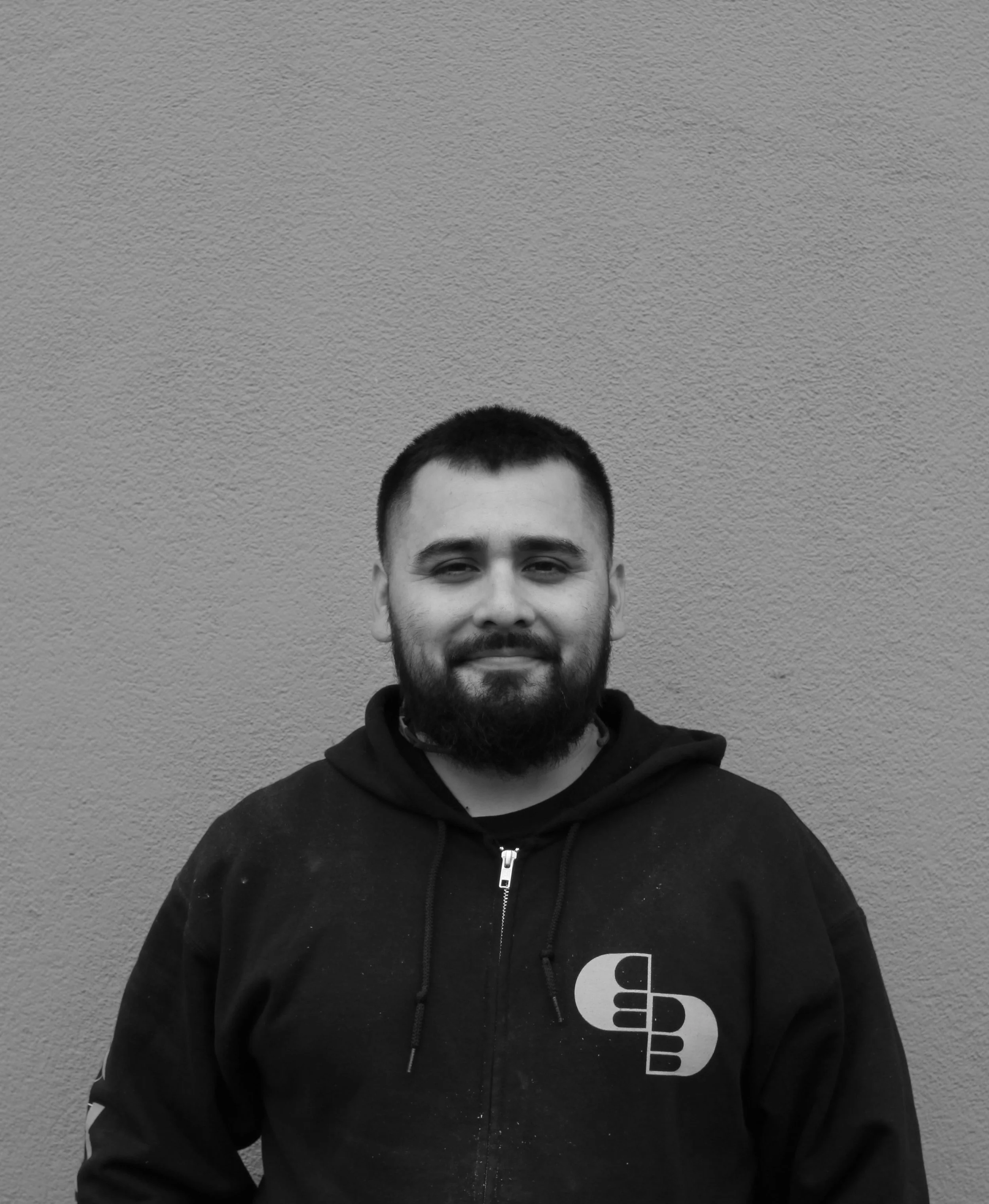A young man with a beard and short hair, smiling, wearing a black zip-up hoodie with a logo on the chest, standing in front of a textured plain wall.
