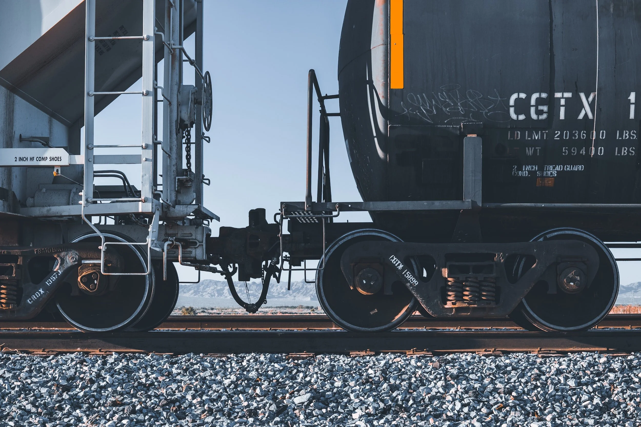 Close-up van een treinwagonsysteem, met zwarte en metalen onderdelen, op smalle spoorrails, met een berglandschap op de achtergrond.