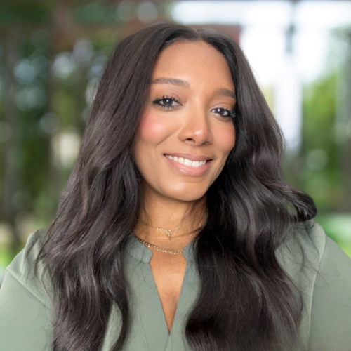 Portrait of a young woman with long dark wavy hair smiling, wearing a light green blouse and gold jewelry, in an outdoor setting with blurred greenery in the background.