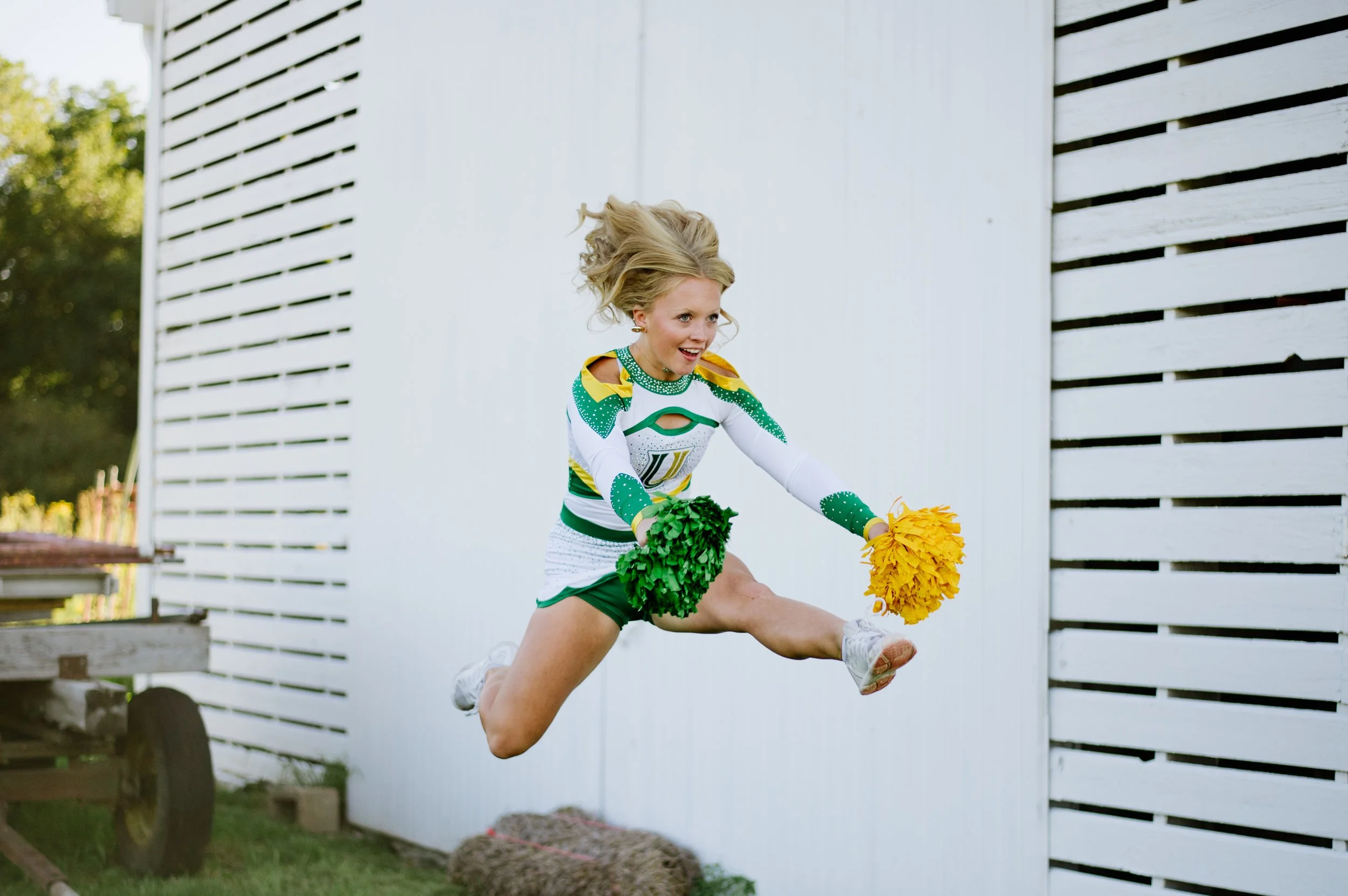 Female cheerleader jumping with pom-poms outdoors near a white wooden wall, wearing a green and white uniform.