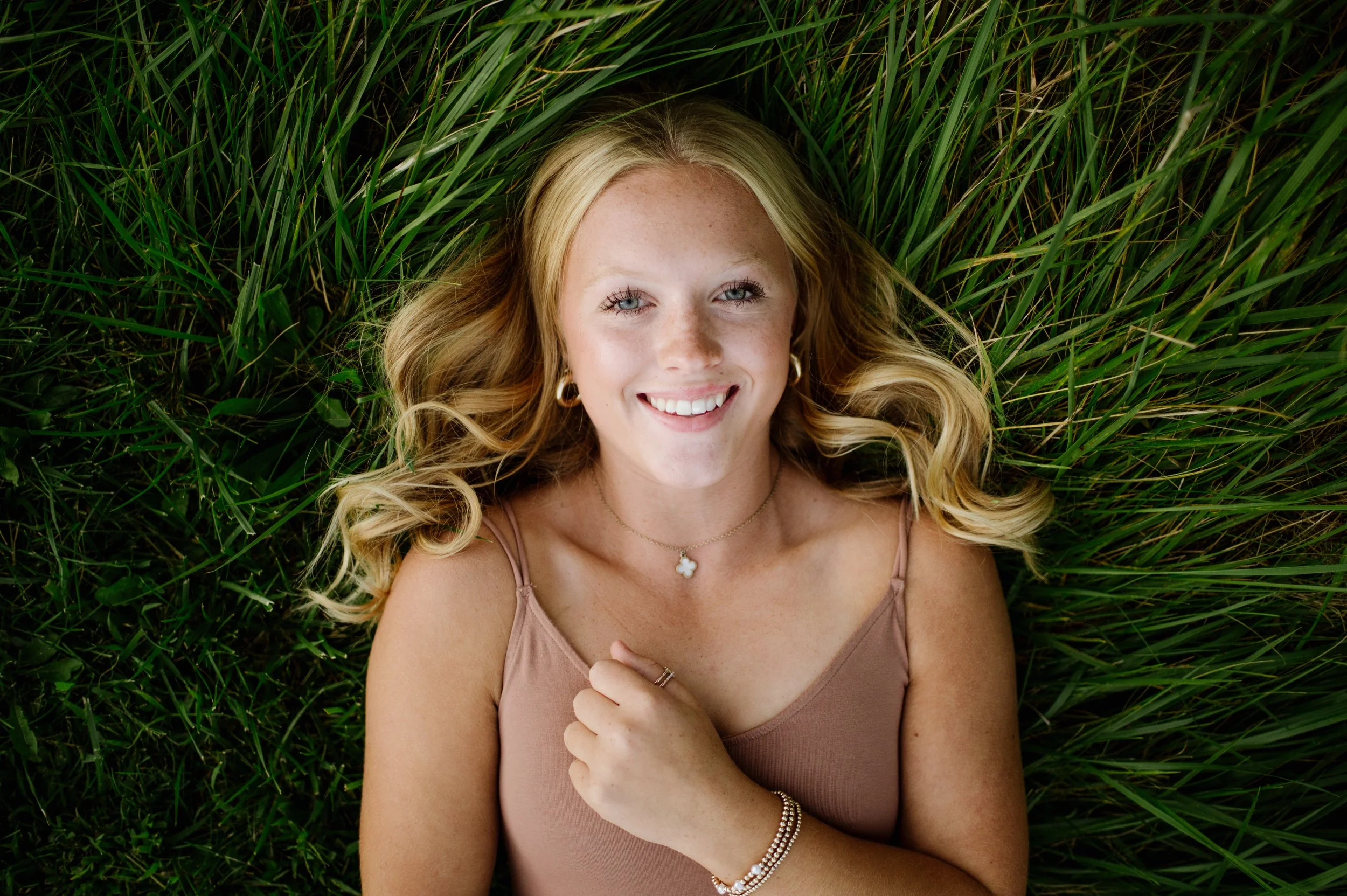A young woman with blond hair lying on green grass, smiling at the camera, wearing a tan tank top, jewelry including earrings, a necklace, and bracelets.