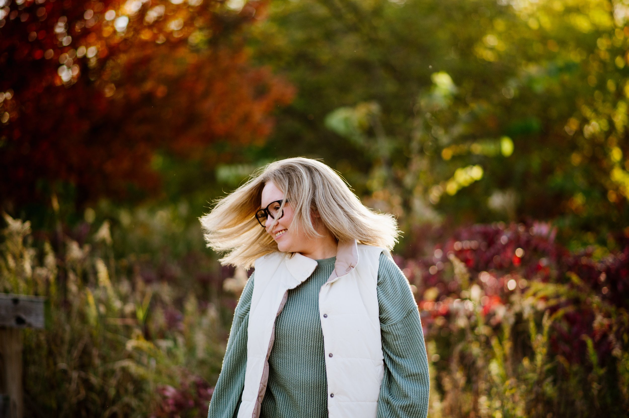 A smiling woman with blonde hair, glasses, wearing a green sweater and a white vest, standing outdoors with colorful fall foliage in the background.