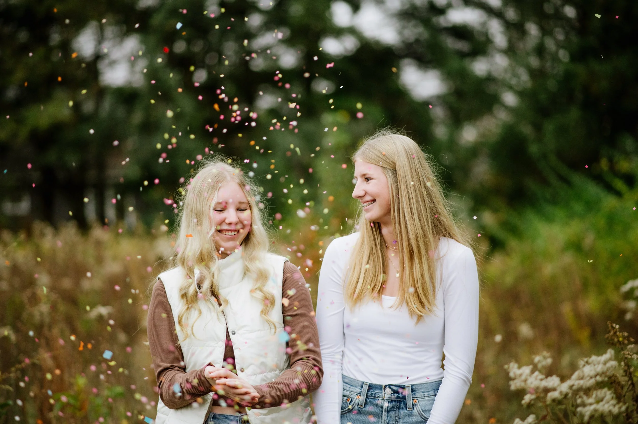 Two women standing outdoors with colorful confetti falling around them, greenery in the background, one smiling with eyes closed and the other smiling at her.