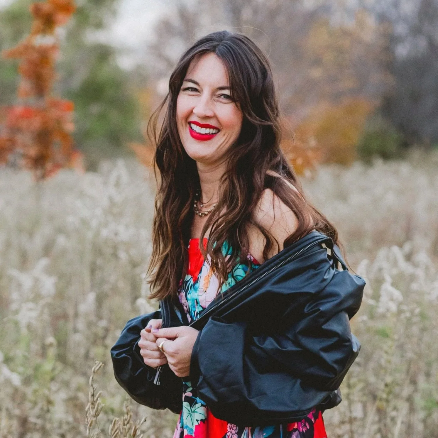 A woman with long dark hair and bright red lipstick smiling outdoors, wearing a colorful floral dress and a black jacket.