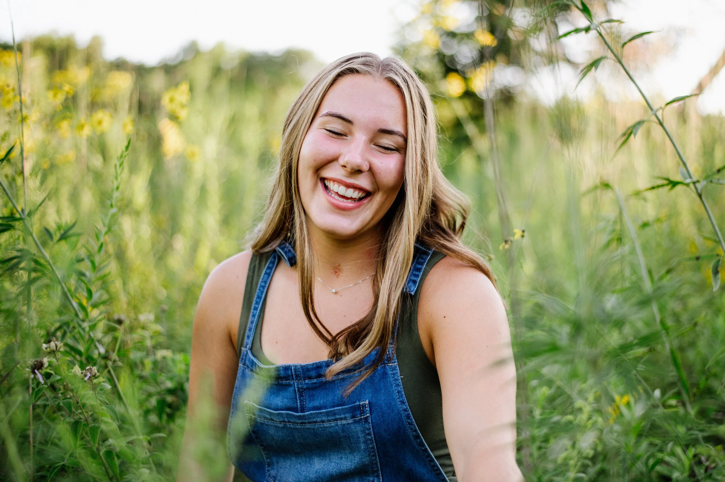 A young woman with long light brown hair and closed eyes smiling broadly, standing in a green field with tall plants and yellow flowers, during daylight.