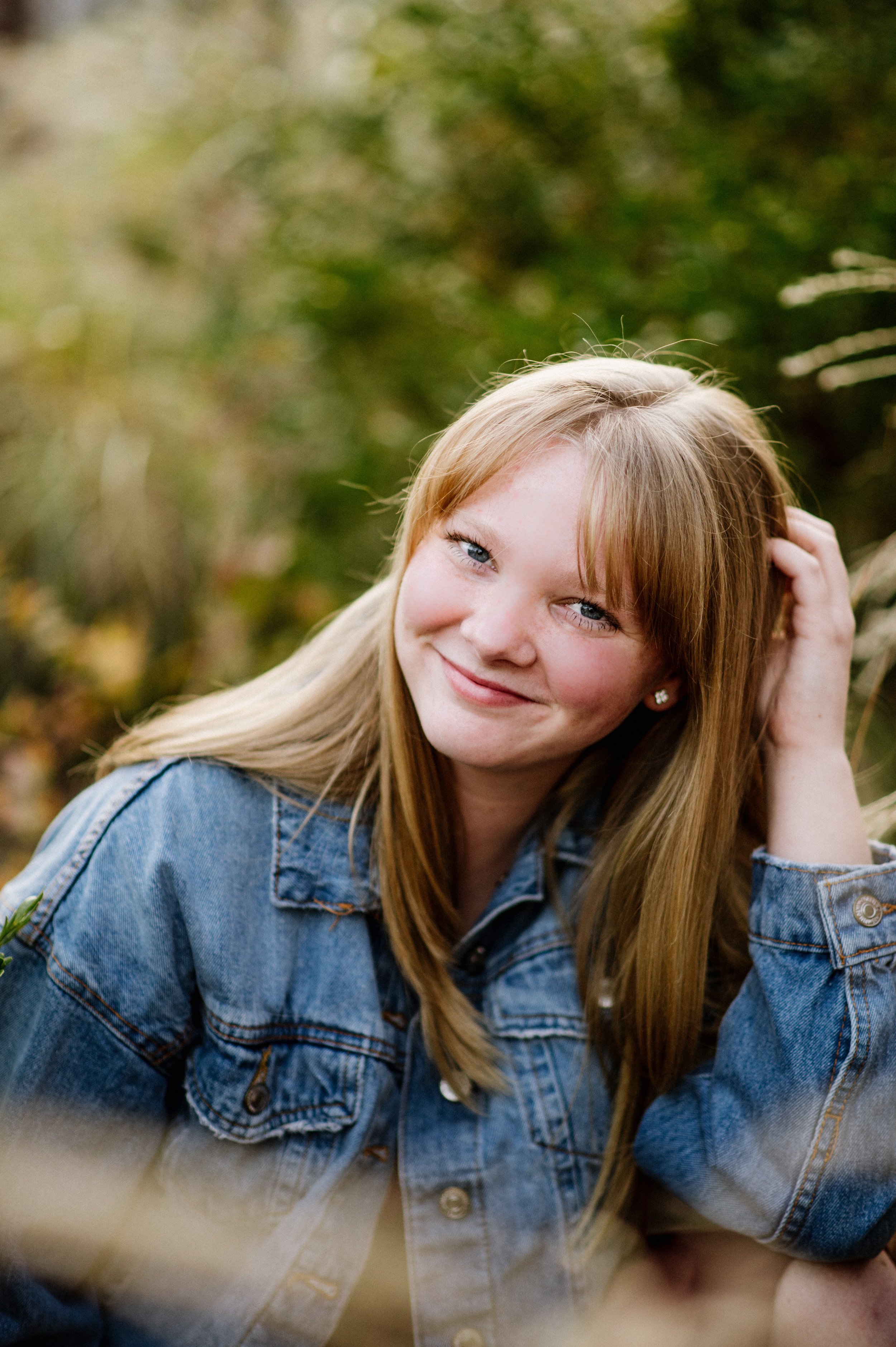 A young woman with long red hair and blue eyes smiling at the camera, sitting outdoors in a natural setting with green foliage in the background, wearing a denim jacket.