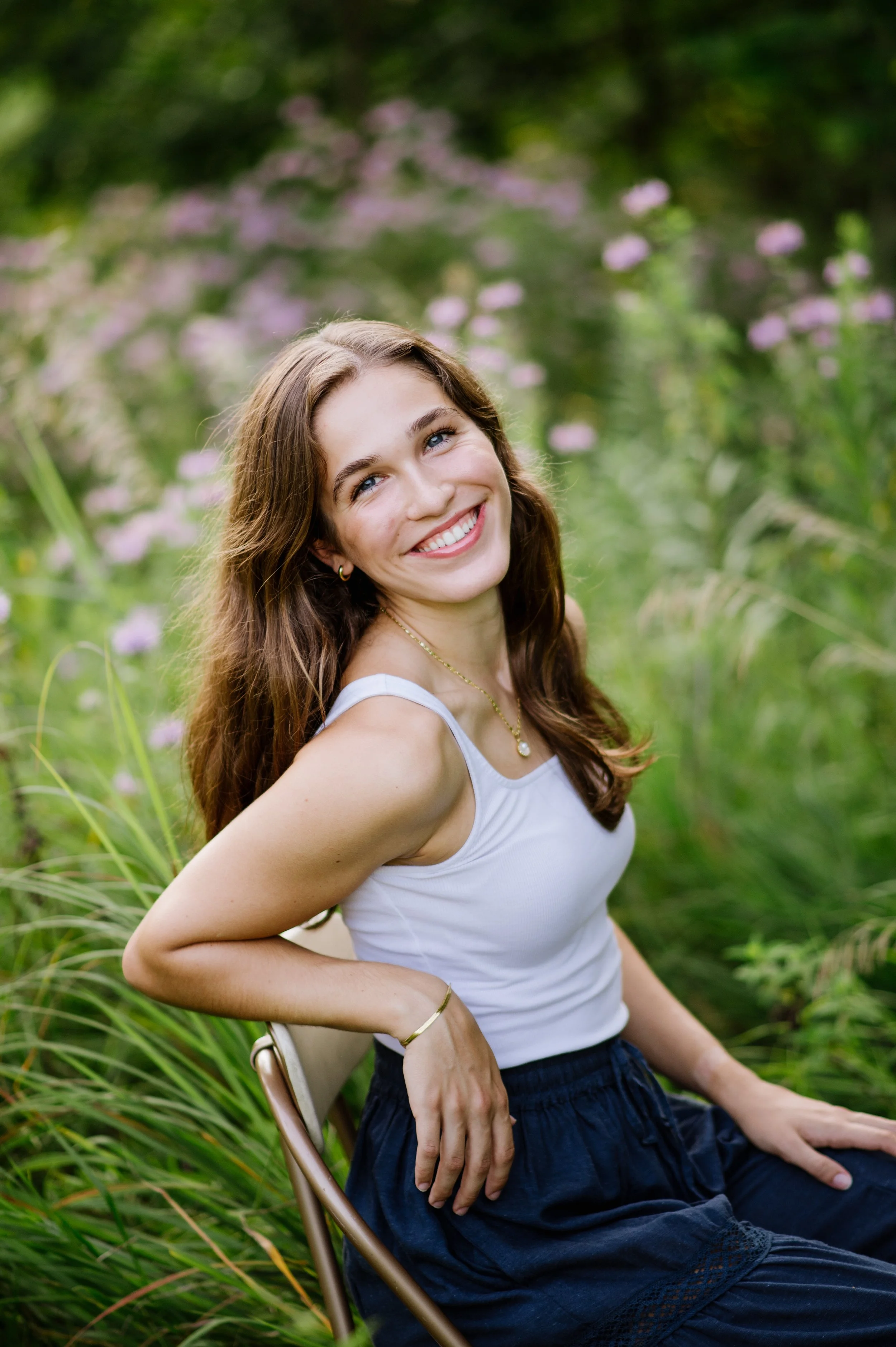 A young woman with long brown hair smiling and sitting outdoors in a lush garden filled with pink flowers and green foliage, wearing a white sleeveless top and navy blue skirt.