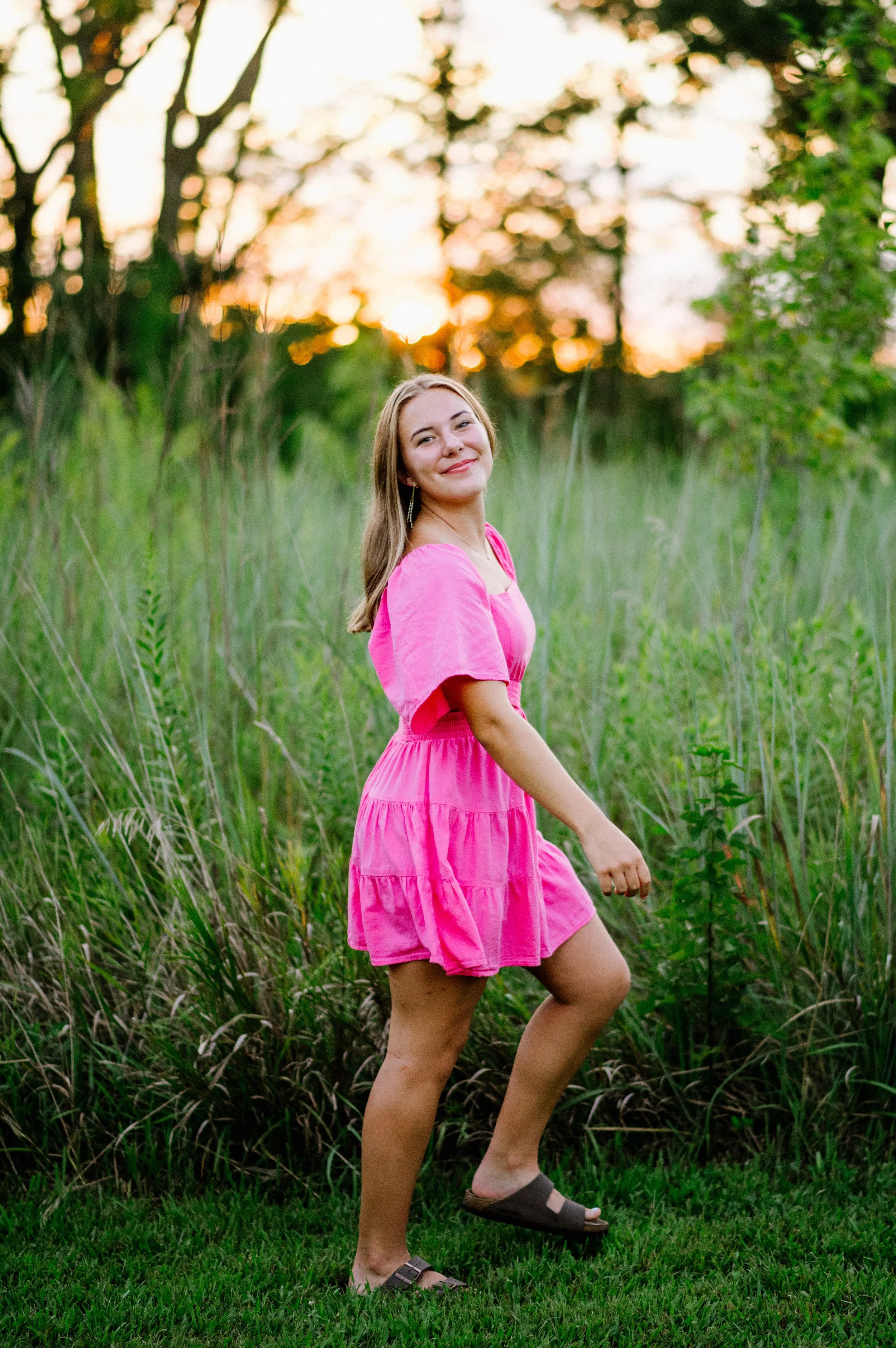 A woman in a bright pink dress standing outdoors in a grassy area surrounded by tall grasses and trees at sunset.
