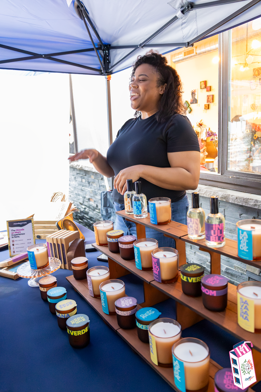 A woman at a display of colorful candles and perfumes under a blue canopy at an outdoor market.