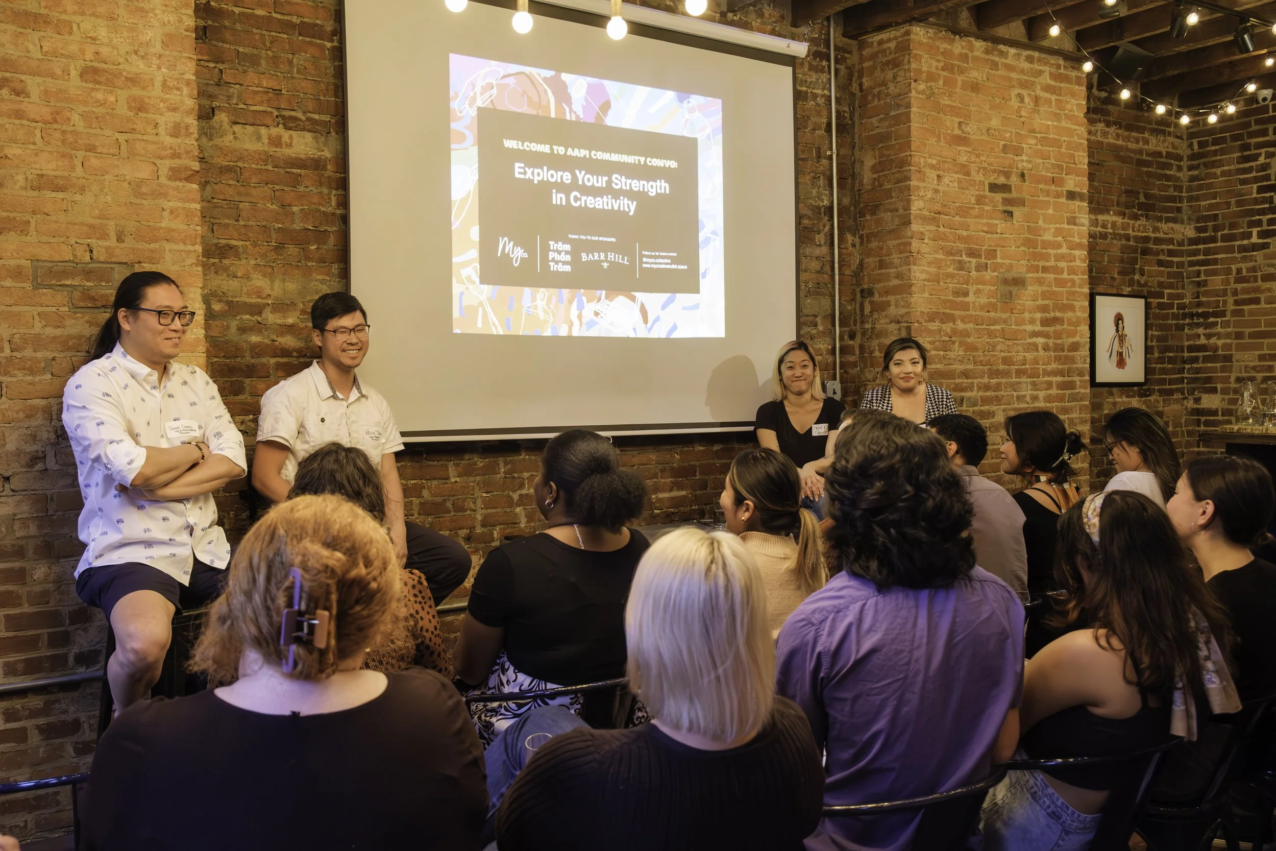 A group of five individuals presenting in front of a seated audience in a brick-walled room with a projector screen. The screen displays a slide titled 'Explore Your Strength in Creativity' at an event called AAPI Community Conversation.