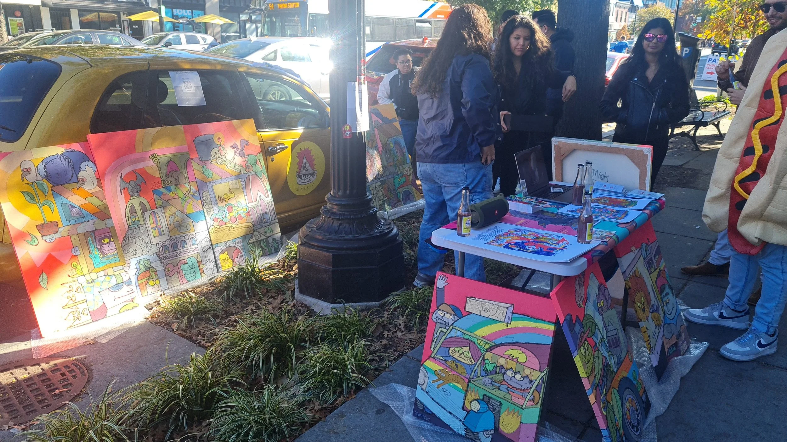 Art vendor's outdoor display with colorful paintings on easels and tables, showcasing cartoon-style artwork of animals and characters, set up on a city sidewalk near a streetlamp, with people browsing and walking past.