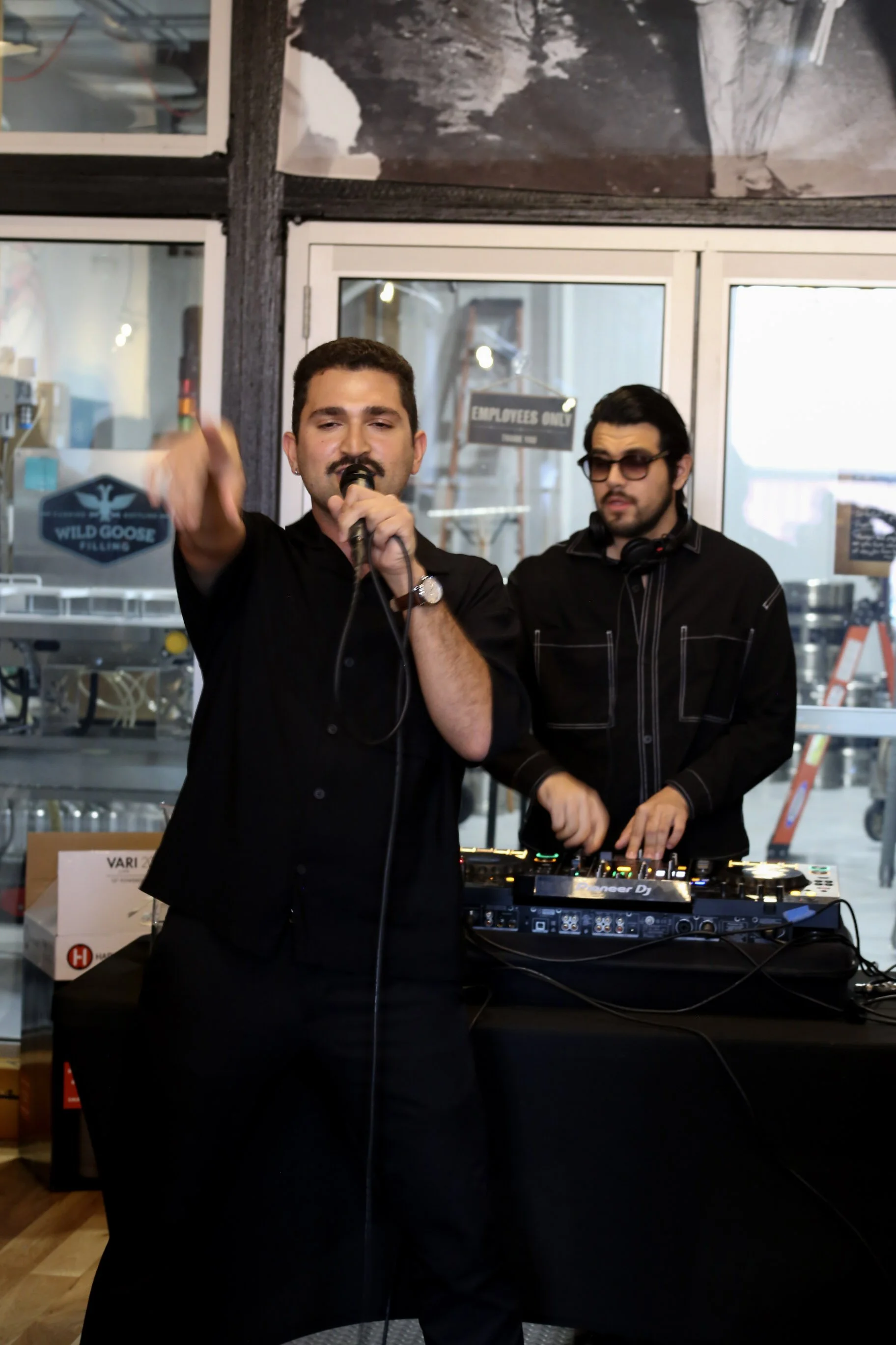 Two men performing music at an indoor venue, one singing into a microphone and the other operating DJ equipment, with signs and a window in the background.