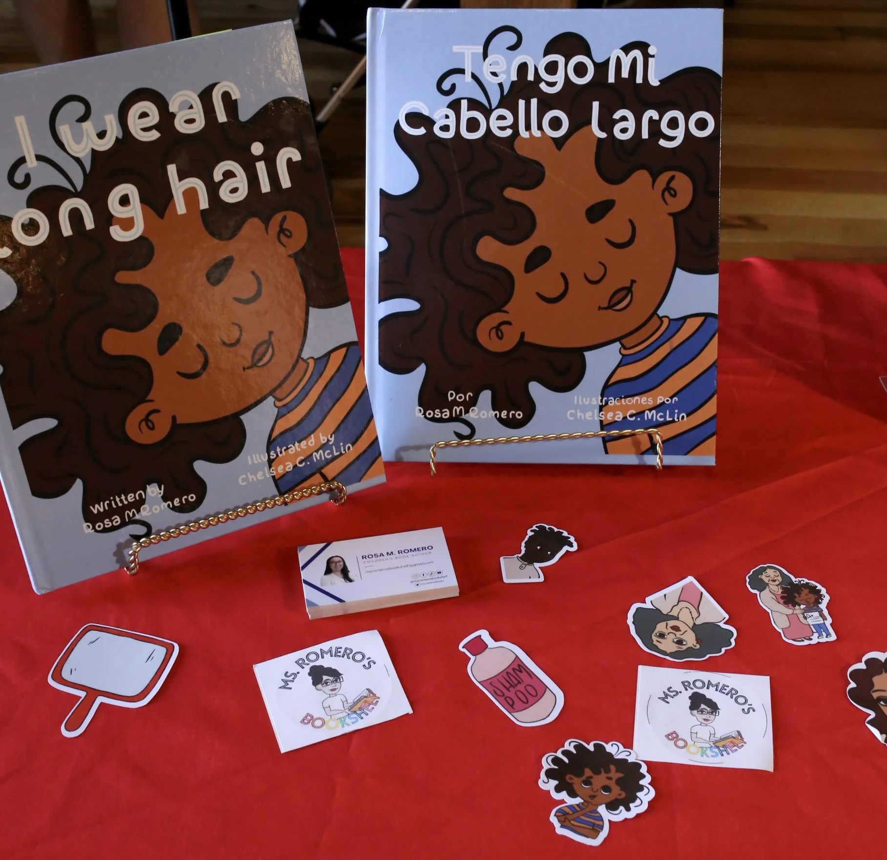 Two children's books with illustrations of a girl with curly hair on display on a table with a red tablecloth, along with stickers, business cards, and small illustrations related to the books.