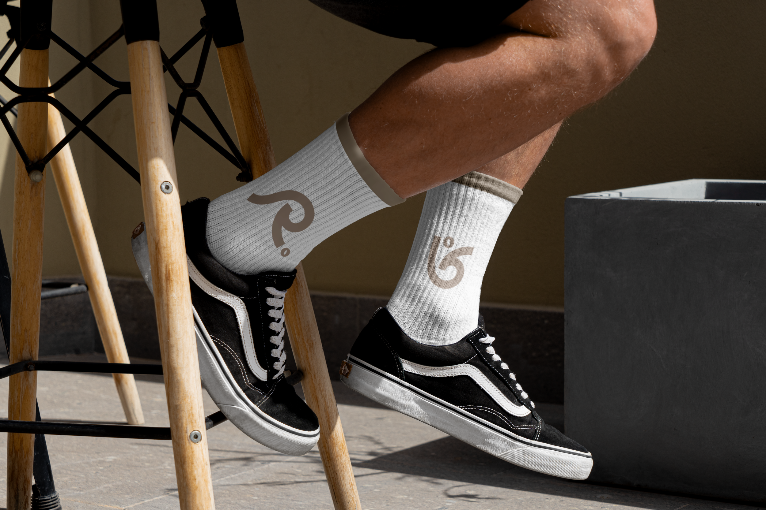 Person wearing black and white Vans sneakers and white socks with logo, sitting on a wooden stool or chair, near a gray planter, with part of their legs visible.