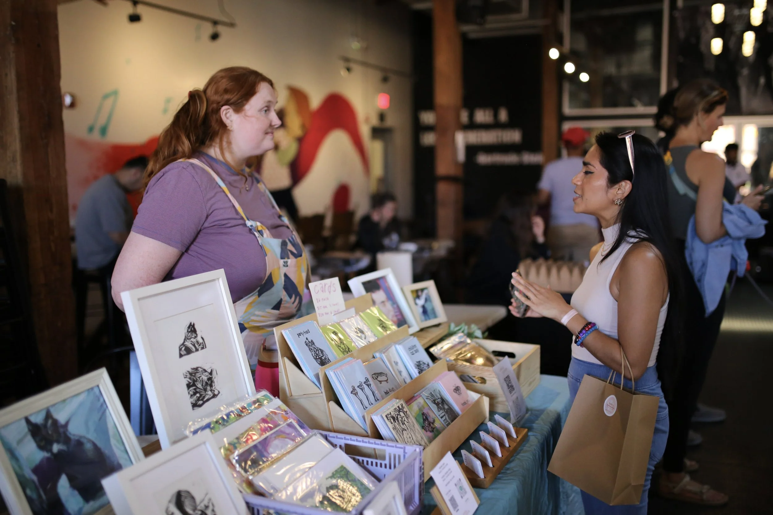 A woman with long black hair, wearing a white sleeveless top, is shopping at a booth with various greeting cards and artwork, speaking with a seller with red hair and a colorful apron in a lively indoor market or craft fair.