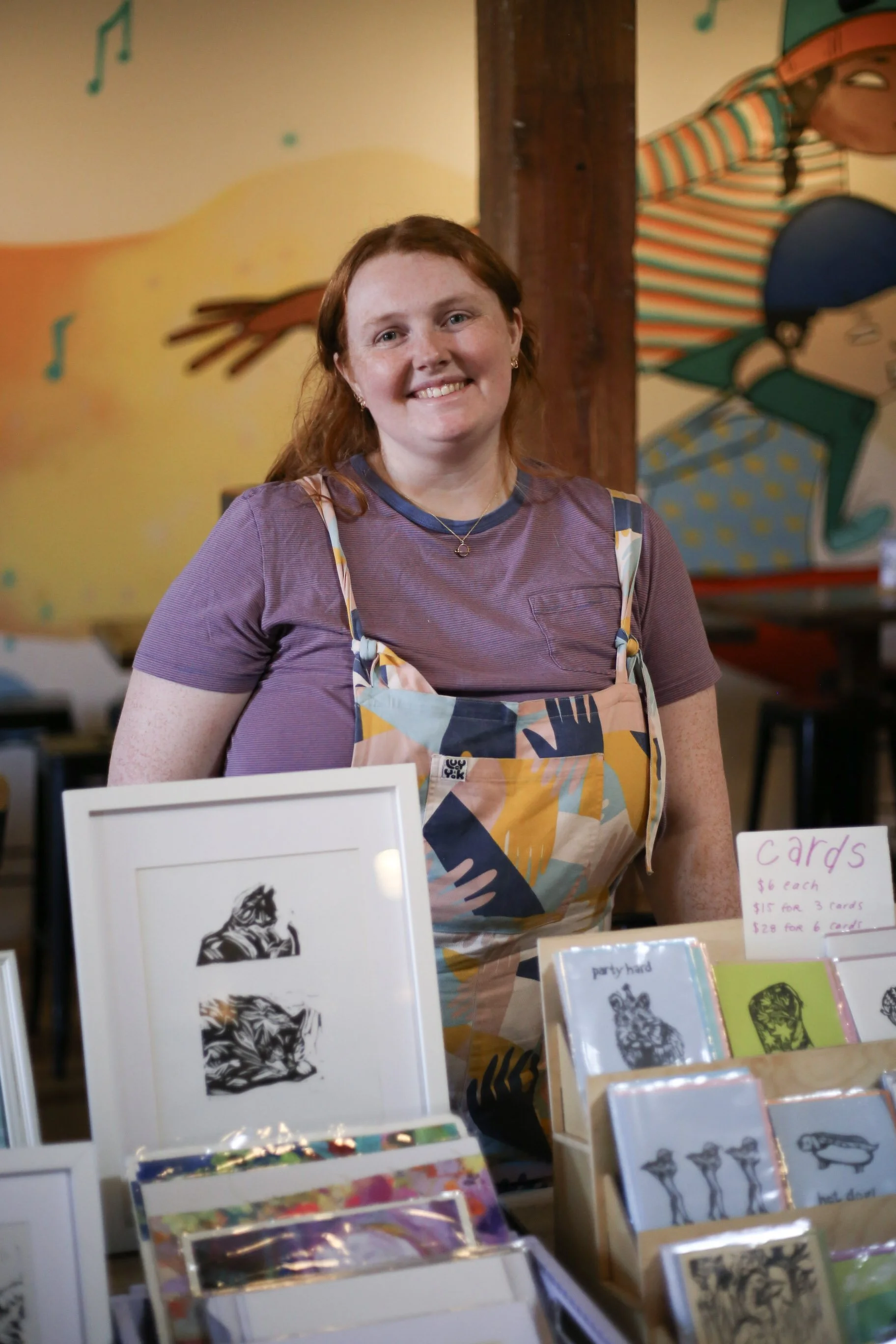 A smiling woman with red hair wearing a purple shirt and a colorful apron at a table selling art and cards at a market or fair.