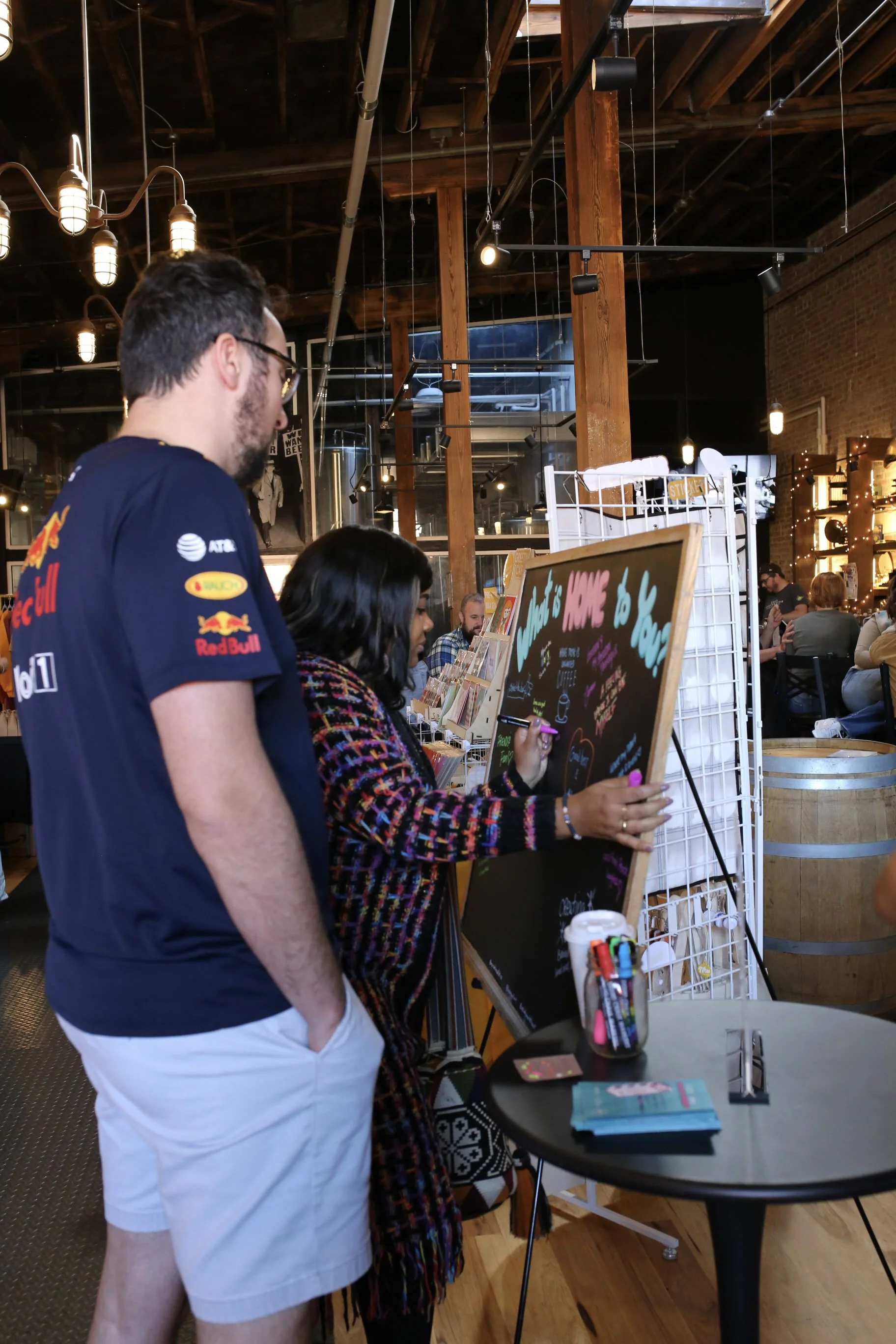 A man in a navy Red Bull racing shirt and white shorts stands next to a woman writing on a black chalkboard sign in a bustling cafe or bookstore with warm lighting and exposed wood beams.