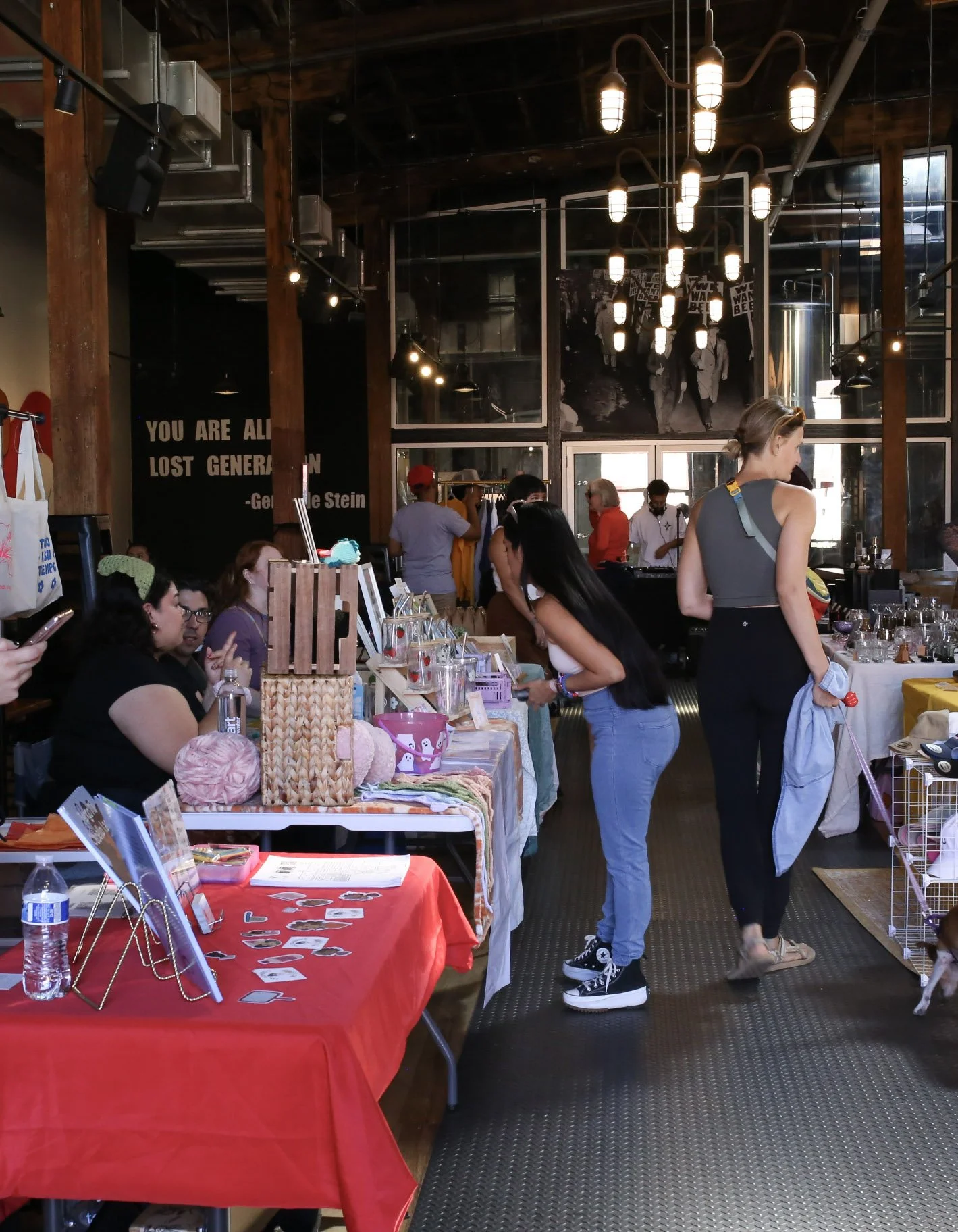 People browsing booths at an indoor market or craft fair inside a large industrial-style building with wooden beams and hanging lights.