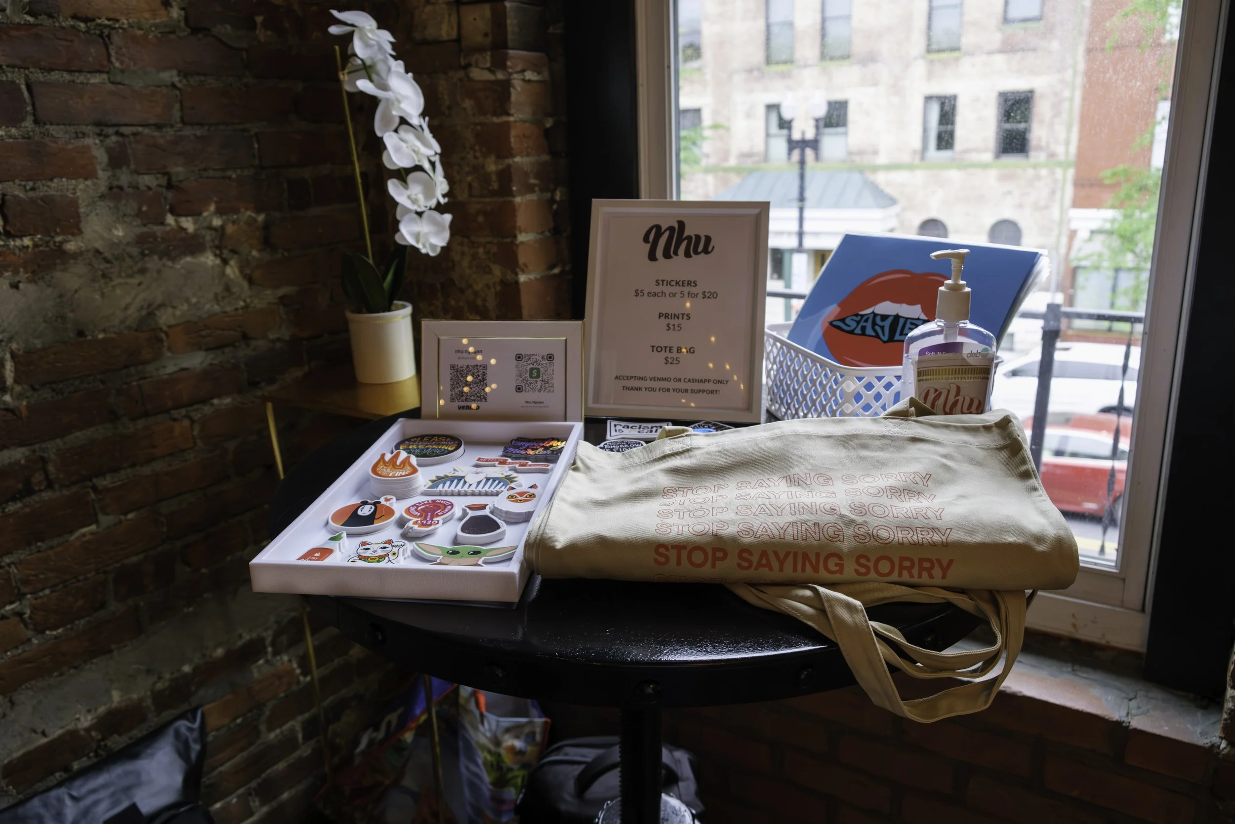 Display of merchandise on a small black table near a brick wall and window. Items include stickers, prints, a tote bag with red text, a bottle of hand sanitizer, and a small white basket with a colorful print. Signs and QR codes are also visible.