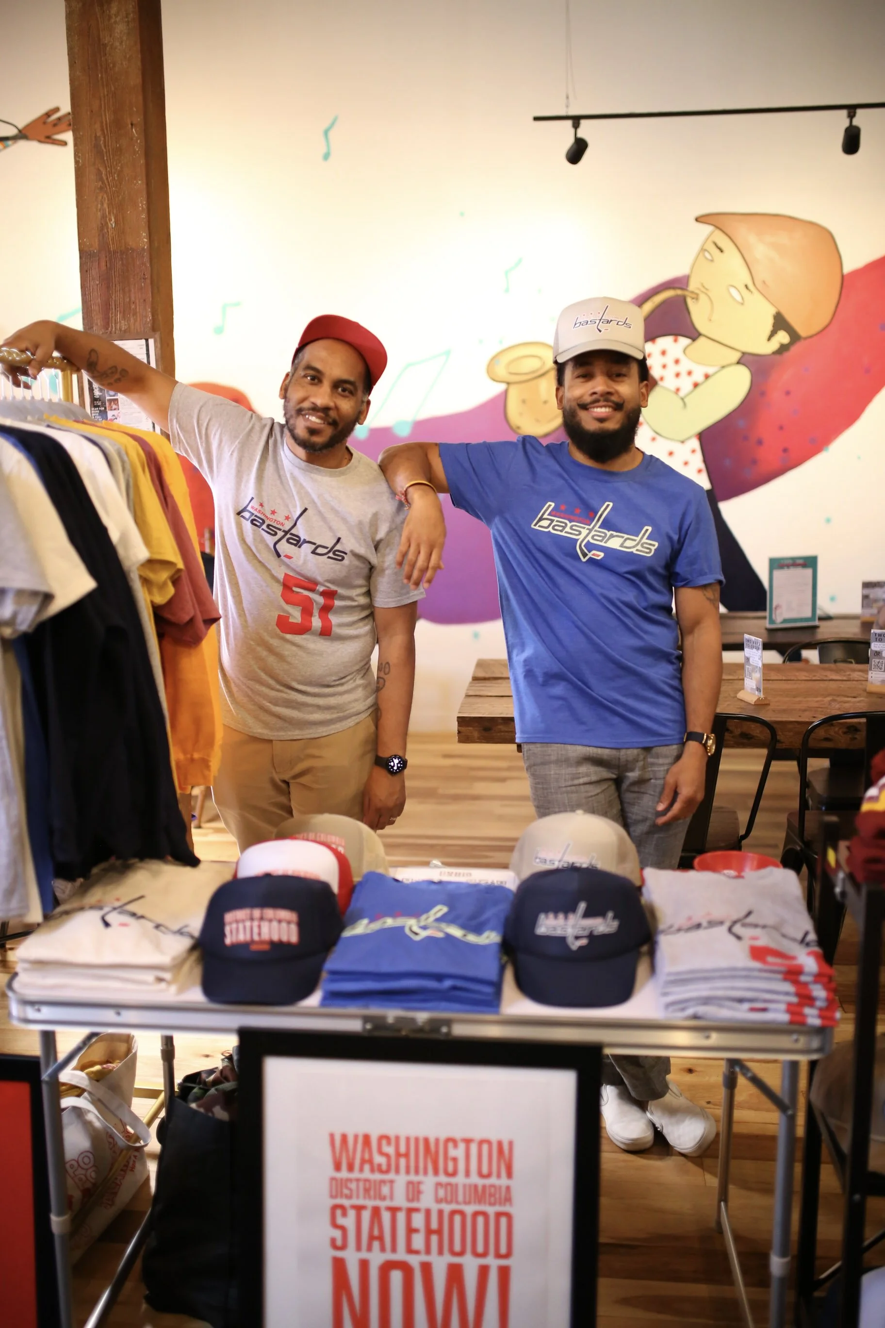 Two men standing behind a table with hats and shirts, smiling for the camera inside a store with a colorful mural background.