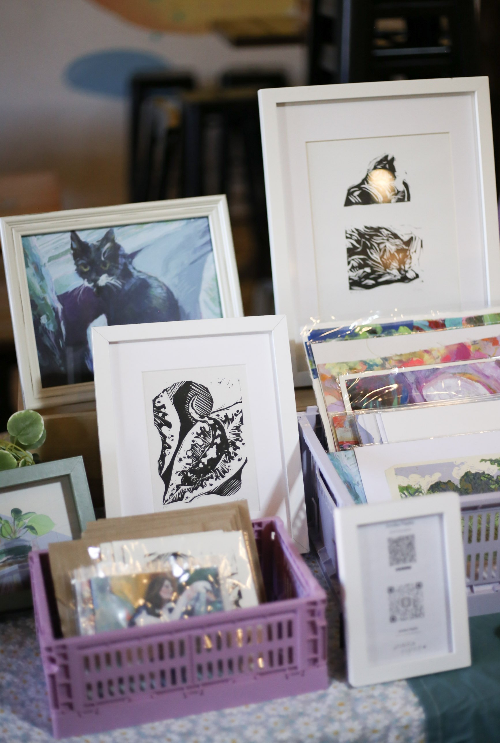 Display of framed artwork and prints on a table at an art fair or market, featuring various images including a portrait of a black cat and black and white sketches.