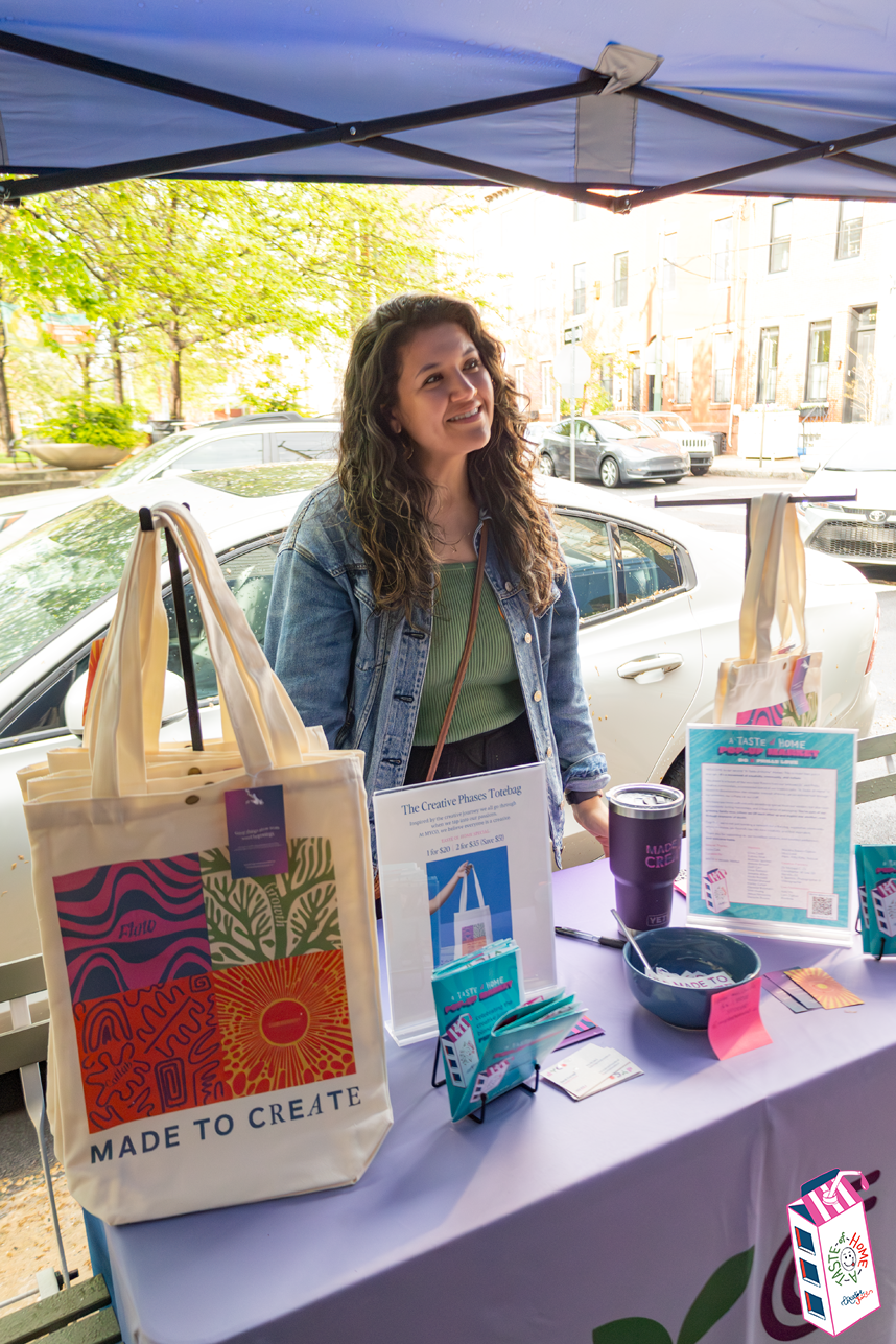 A woman standing at a booth with her hands resting on a table, smiling, surrounded by tote bags and informational signs, outdoors with cars and trees in the background.