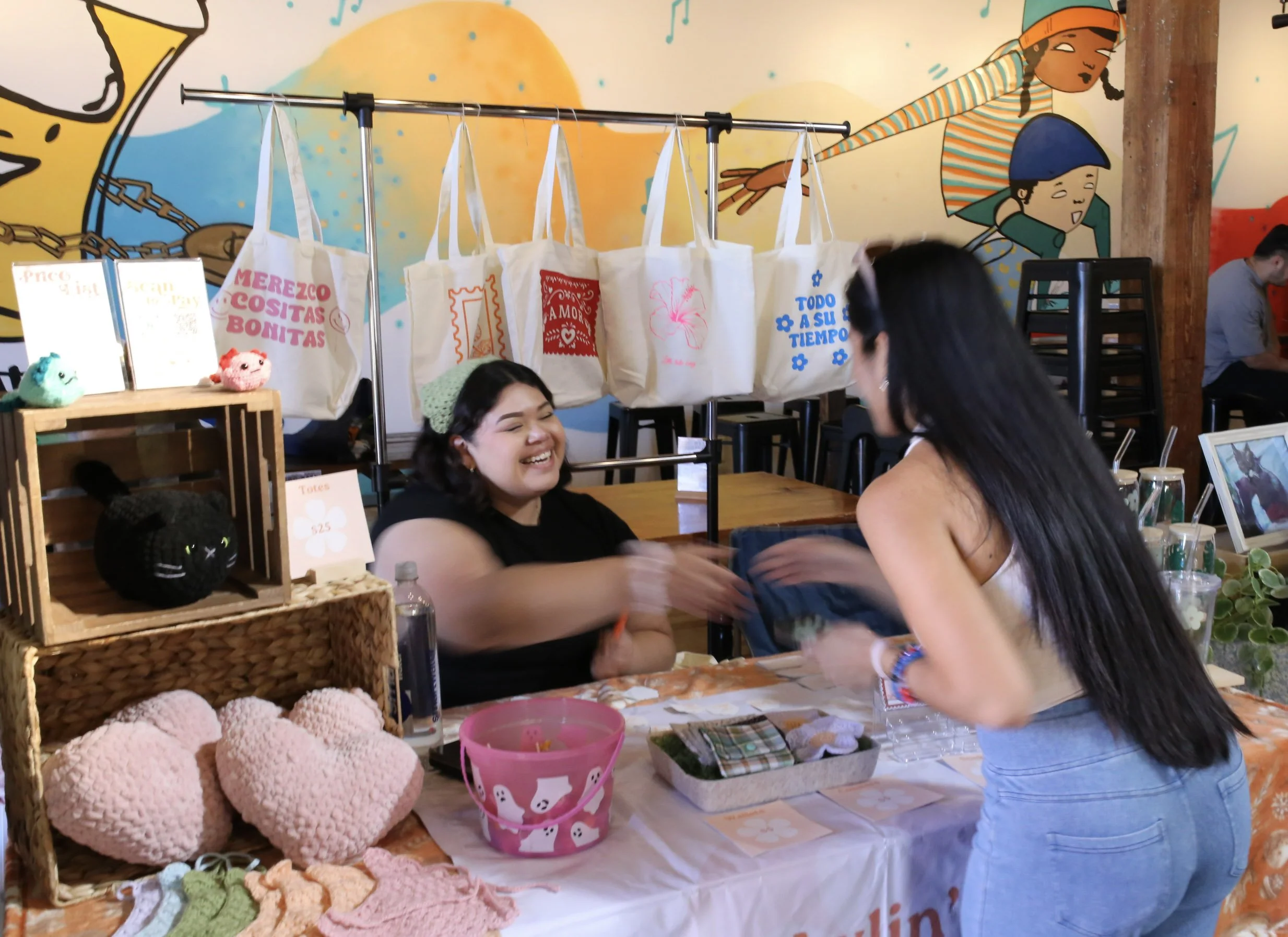 Two women at a craft or gift shop, smiling and shaking hands across the table. Behind the woman on the table, there are handmade stuffed hearts and yarn crafts. The background features colorful wall art and hanging tote bags with various designs and phrases, with some stacked chairs and a person seated reading in the background.
