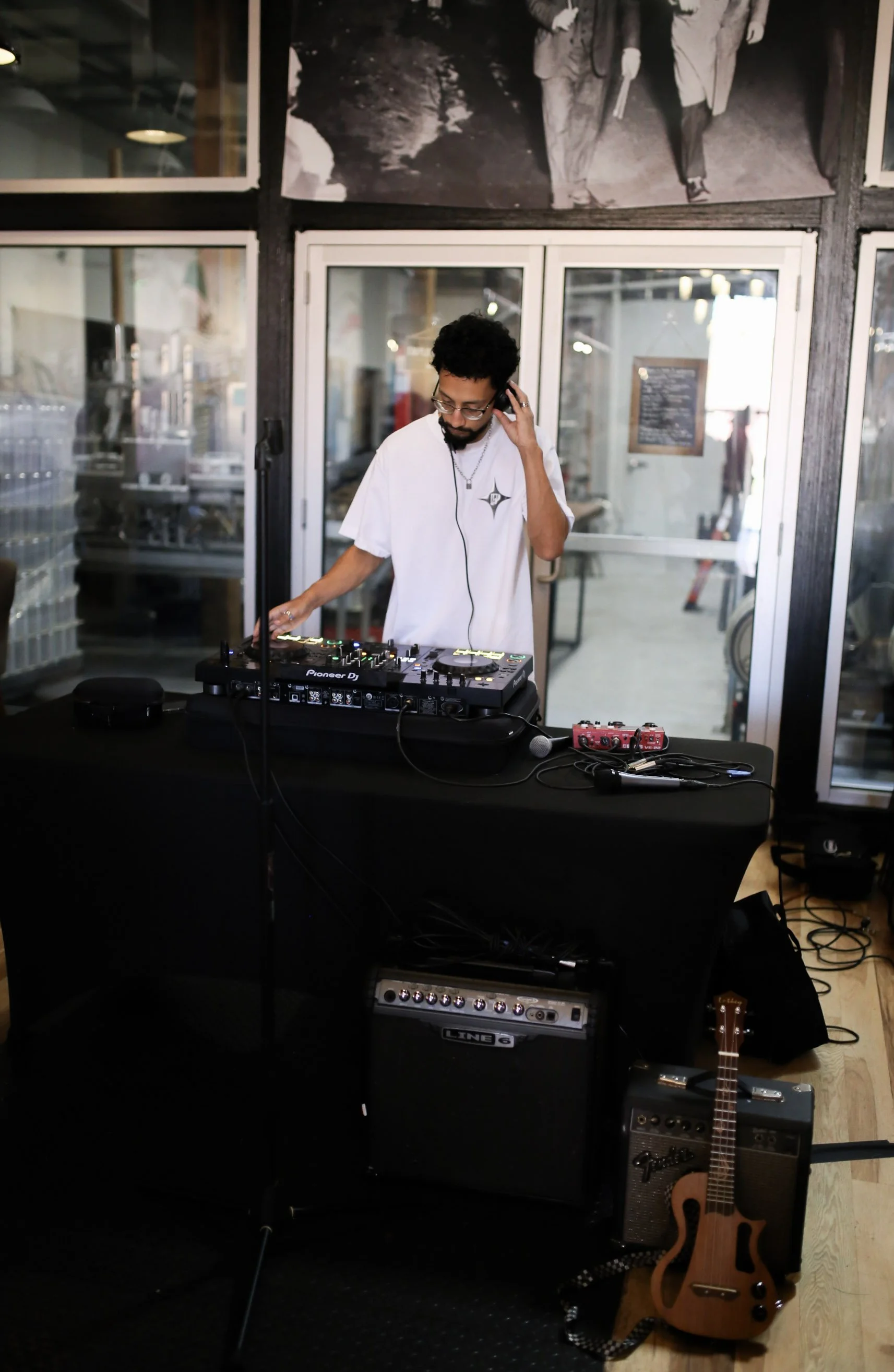 A DJ is performing at an indoor venue, wearing a white T-shirt and headphones, with DJ equipment, a guitar, and amplifiers around him.