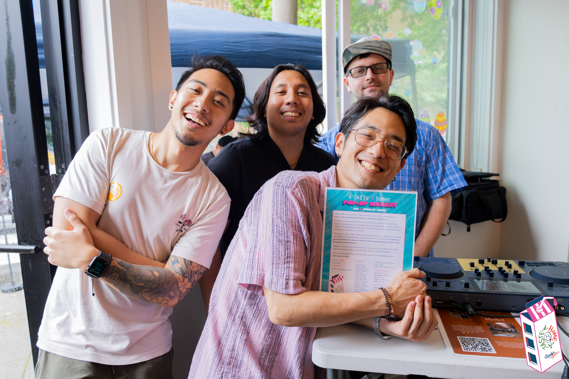 Five smiling people indoors at a community event, with one holding a colorful flyer and a DJ setup nearby, celebrating together.