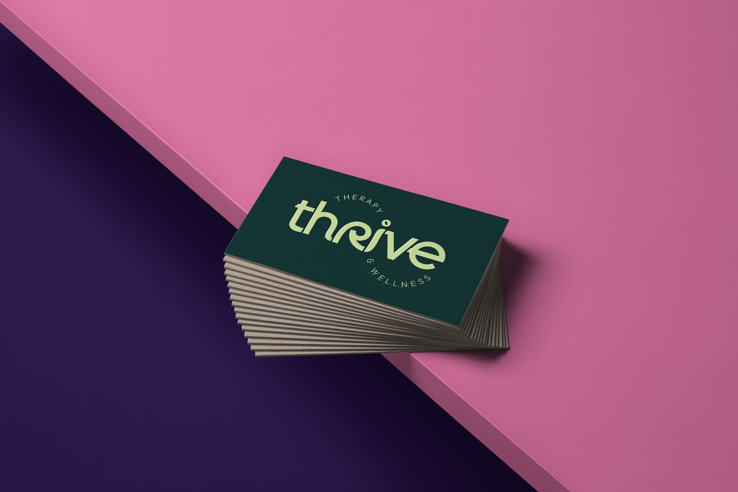 A stack of business cards on a surface divided diagonally with pink and purple colors. The top card displays the word 'thrive' with the words 'therapy' and 'wellness' in a circular arrangement.