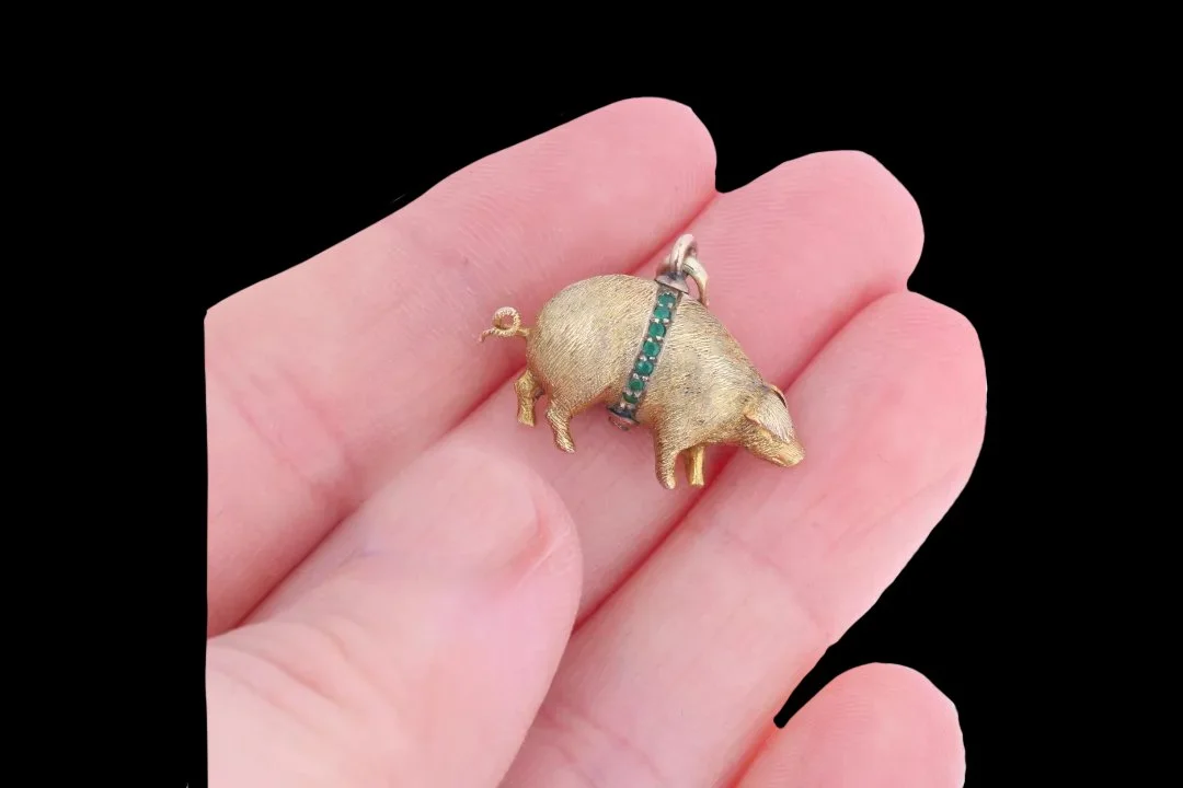 Miniature antique gold lucky pig charm resting on fingertips, showing its 23 mm length and green paste-set decorative band.