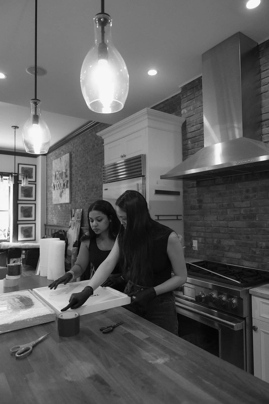 Two women wearing black shirts and black gloves working together at a kitchen counter, preparing ingredients, with kitchen appliances and brick walls in the background.
