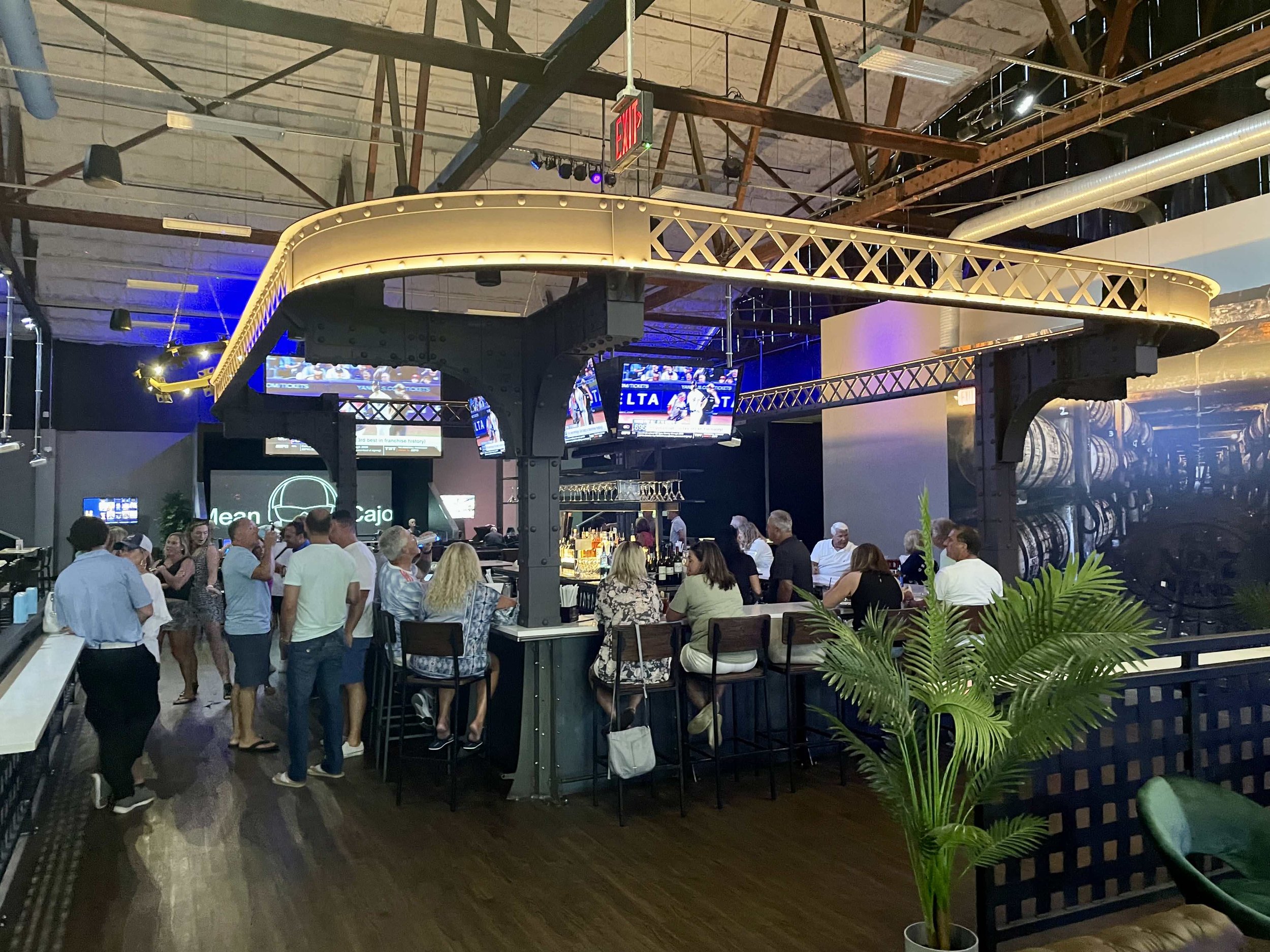 Guests enjoying drinks at the circular bar at Back 9 Social in Wallingford, CT, featuring signature gold bridge-truss overhead lighting and sports on multiple screens