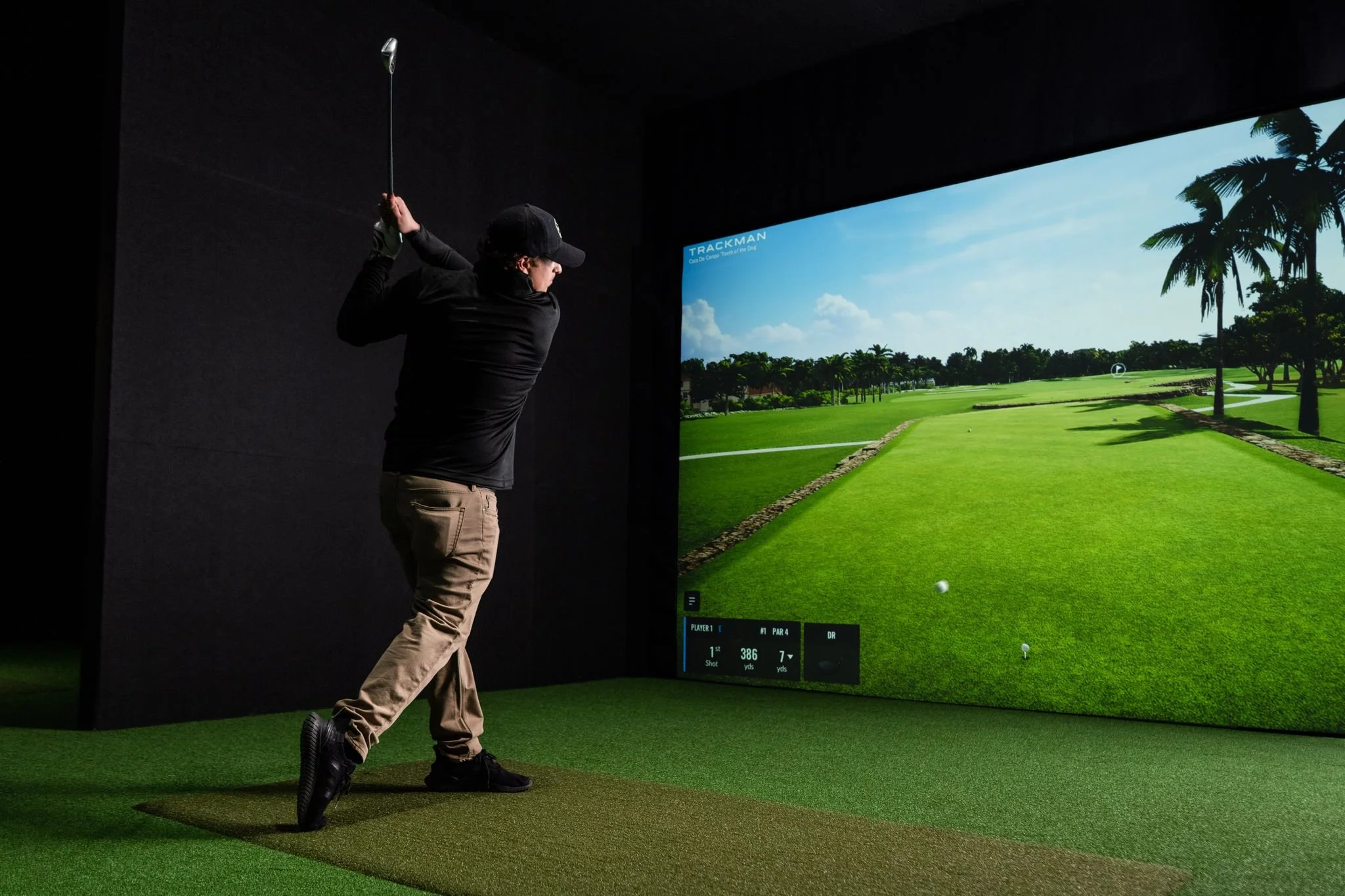 Golfer mid-swing on a Trackman golf simulator at Back 9 Social, with a tropical course displayed on the large projection screen