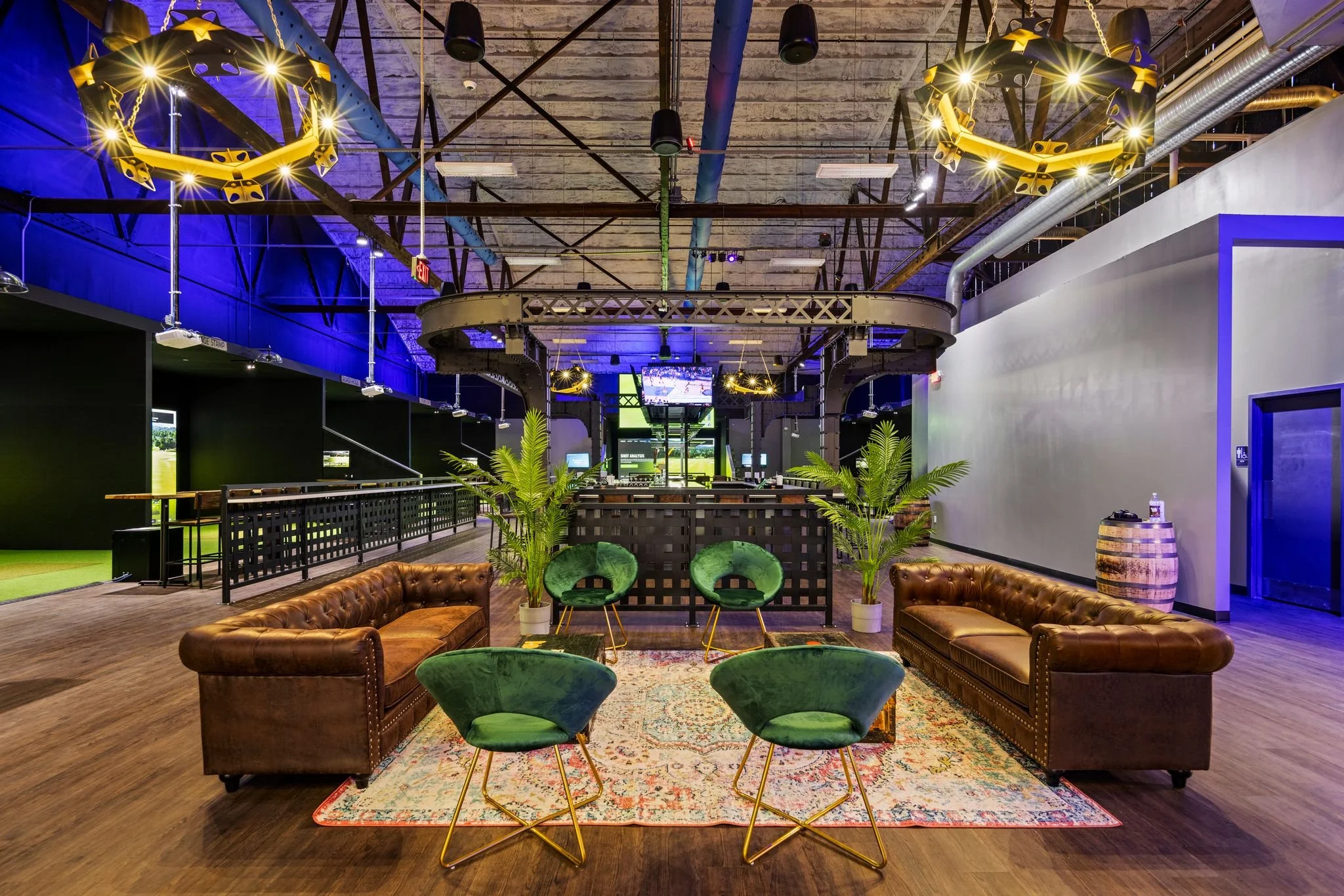 Stylish lounge area at Back 9 Social Wallingford CT with brown leather Chesterfield sofas, green velvet chairs, and a view of the Trackman golf simulator bays and central bar
