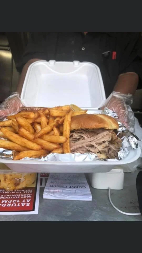Foil-wrapped pulled pork sandwich with French fries in a white foam takeout container, placed on a table with receipts and advertisement flyers.
