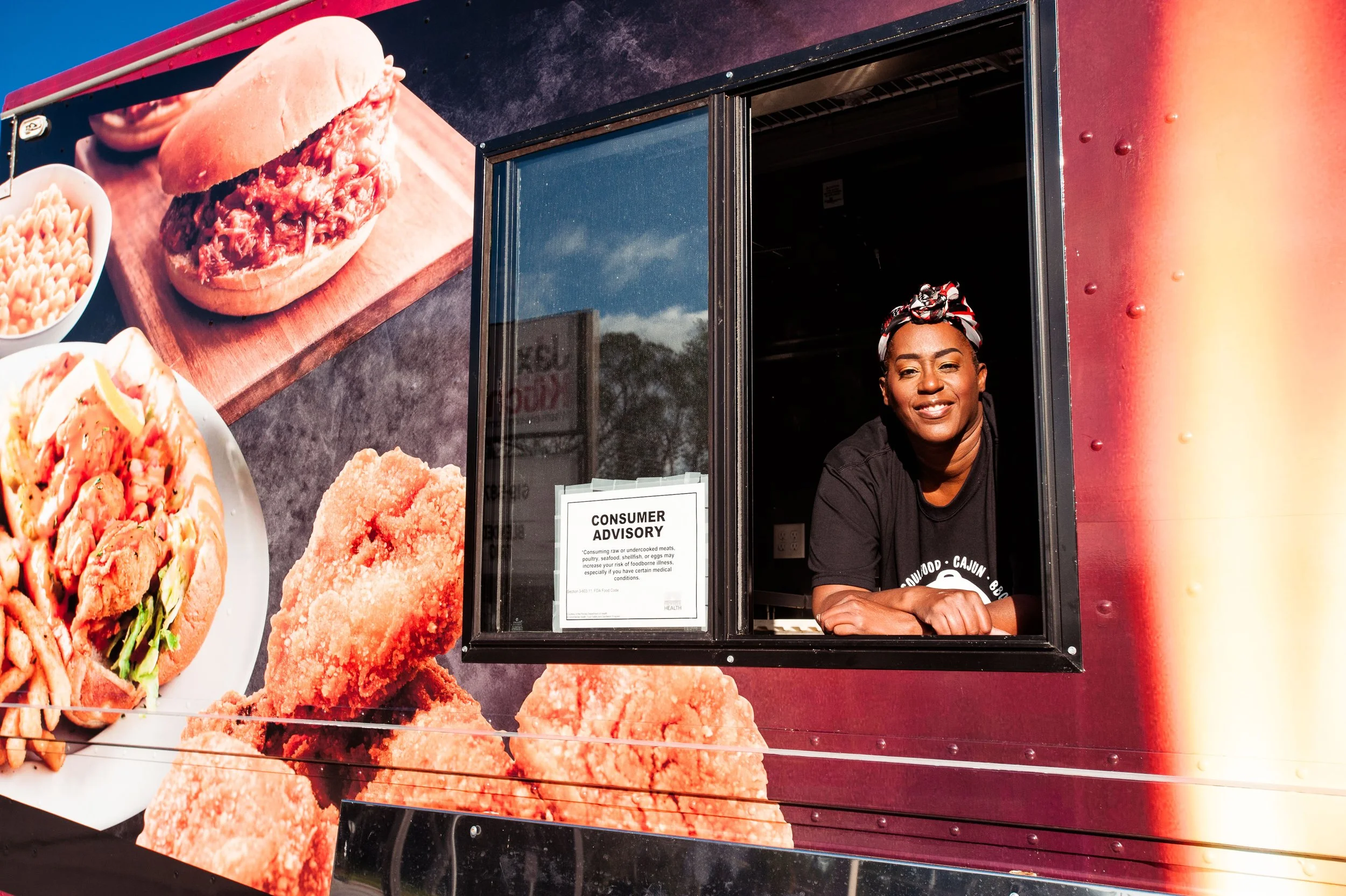 A woman leaning out of a food truck window, smiling. The truck's exterior features large food images including chicken wings, fried chicken, and sandwiches. There is a consumer advisory sign in the window.