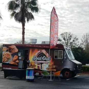 Food truck named 'Soul Out' parked outdoors with a pink promotional flag, a table and chairs, palm trees, and cloudy sky in the background.