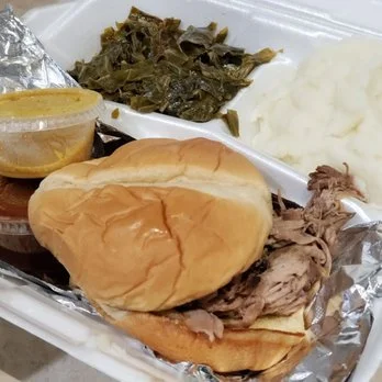 Sandwich with shredded beef, white bread bun, and a side of greens, mashed potatoes, and barbecue sauce