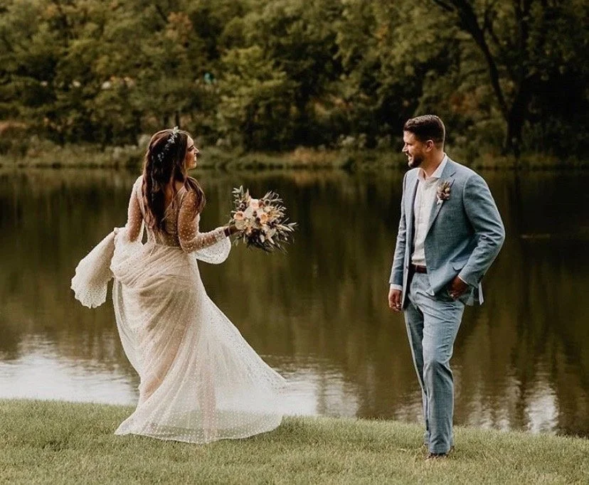 Bride and groom by a pond, bride holding a bouquet, bride in a long wedding dress, groom in a light blue suit, outdoors with trees in the background.