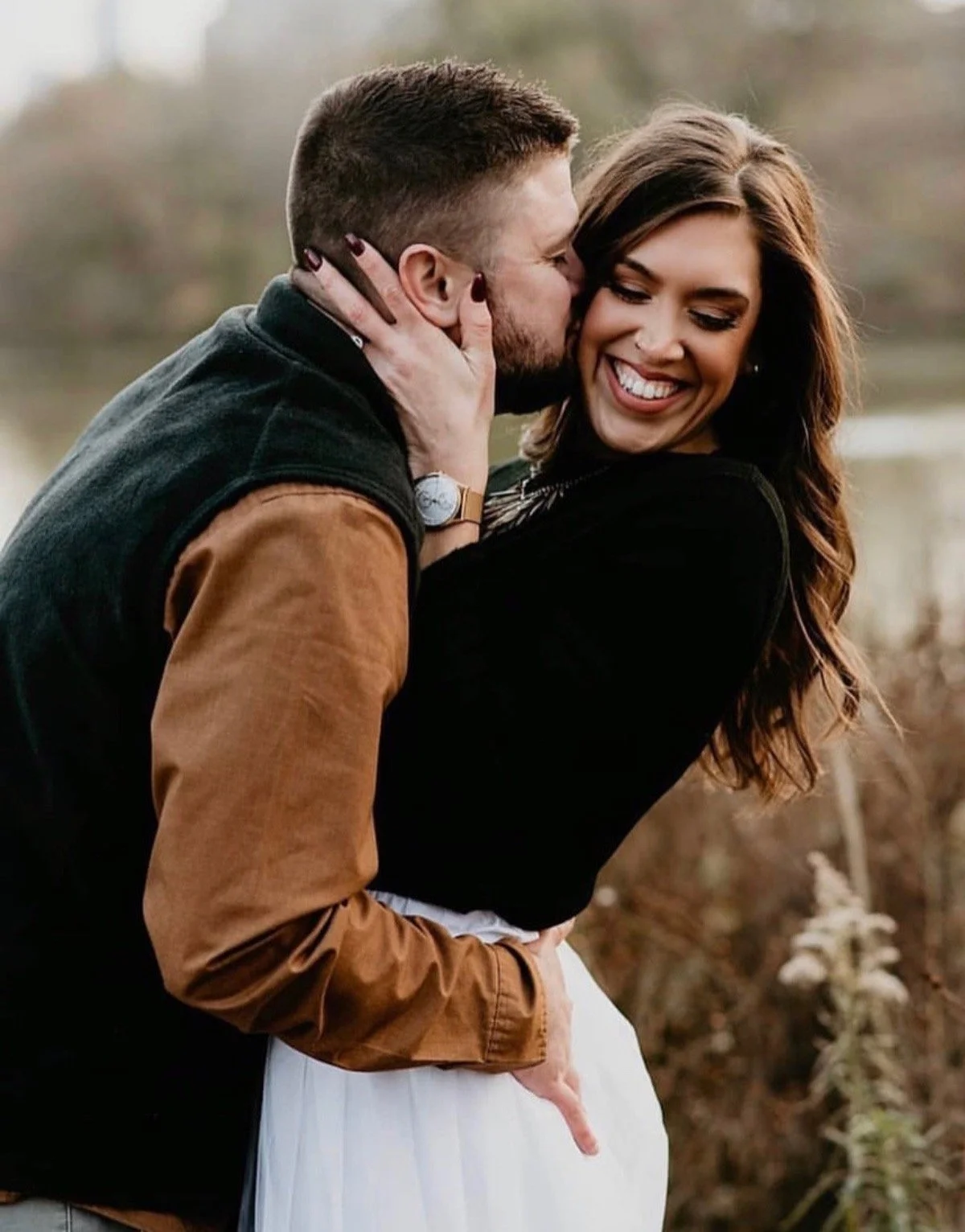A happy couple outdoors, with the man kissing the woman's cheek, both smiling warmly.