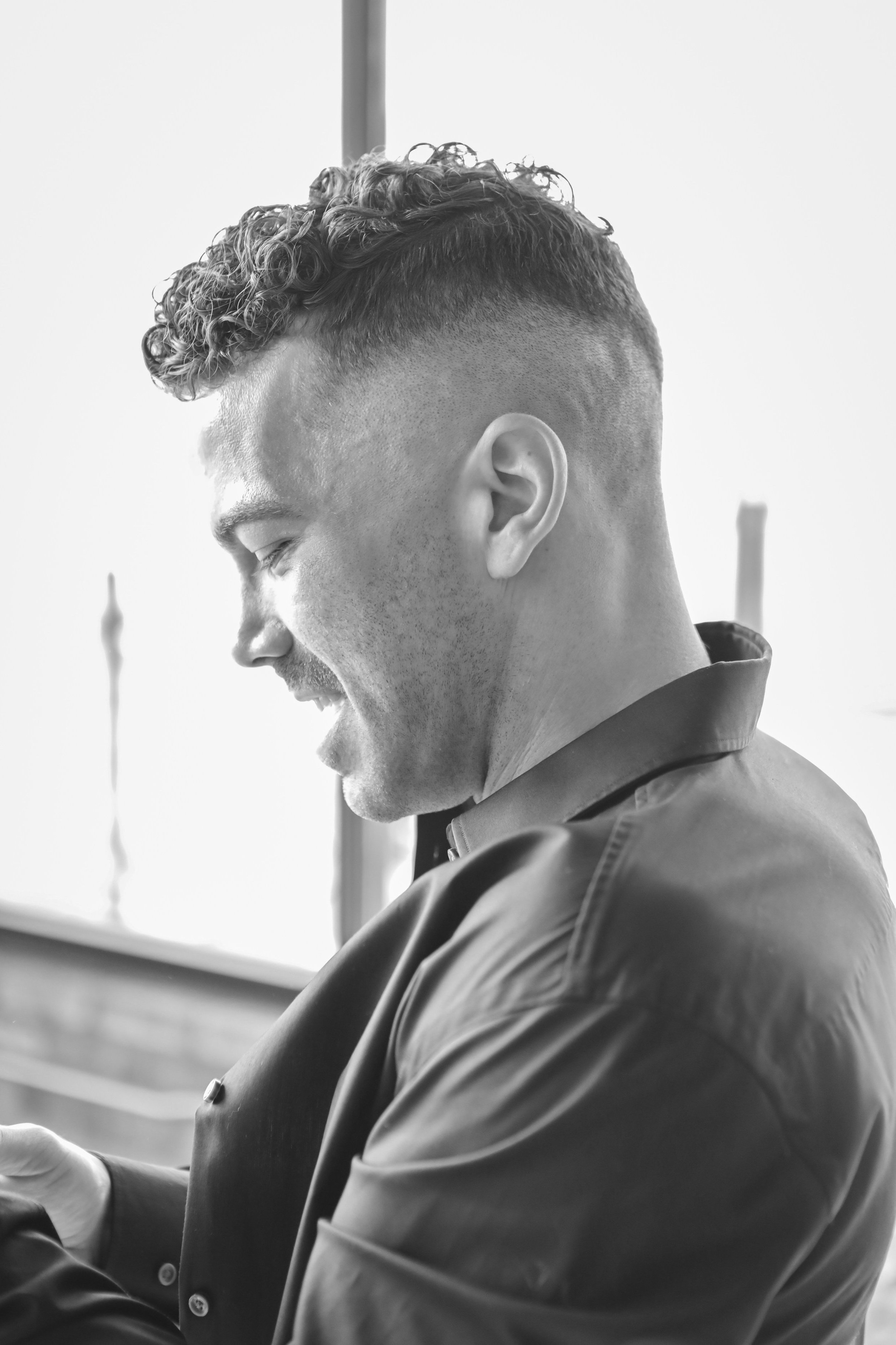 Black and white photo of a young man with curly hair and a fade haircut, smiling and looking downward, wearing a collared shirt and a jacket.