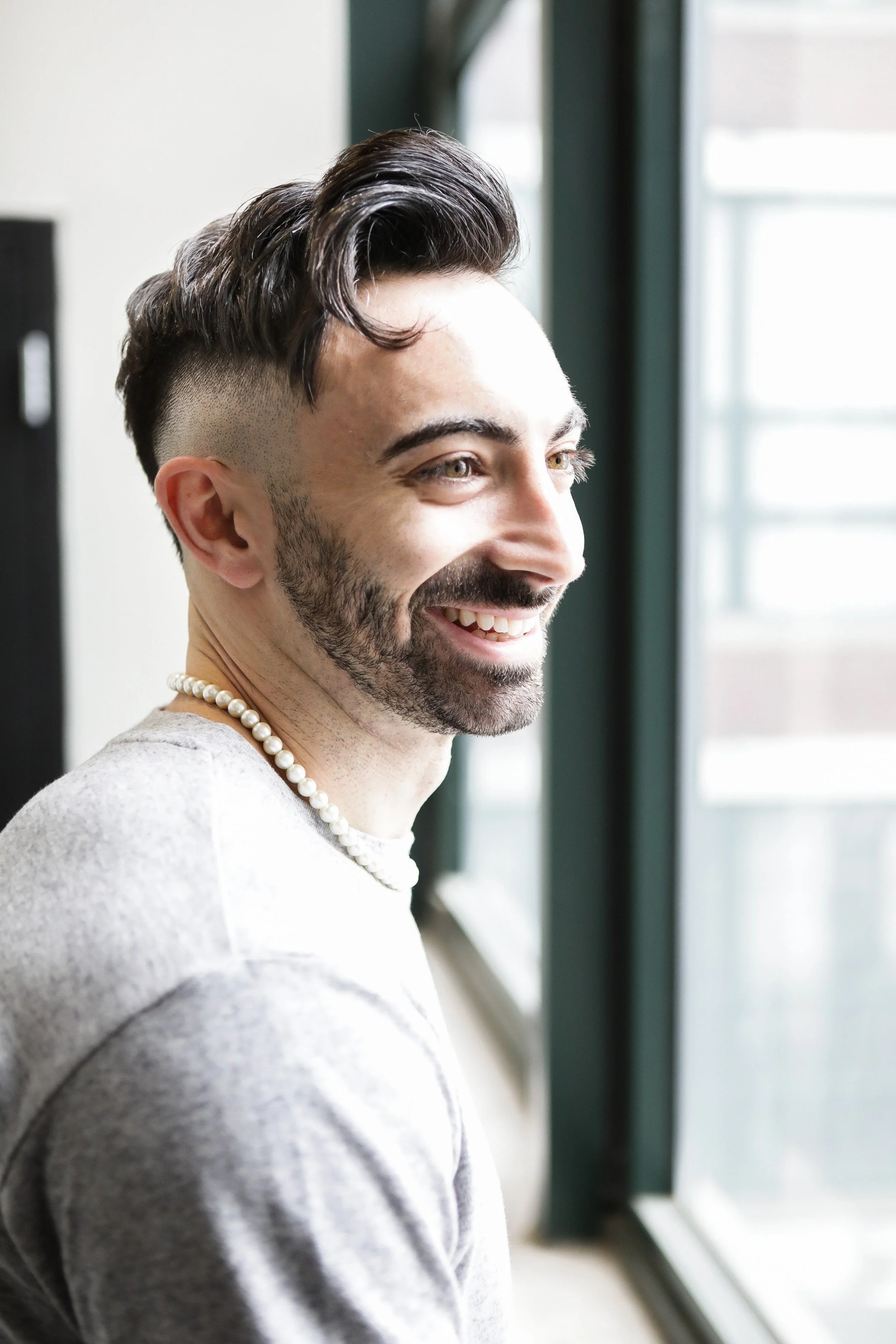 A smiling man with dark hair and a beard looking out a window, wearing a pearl necklace and a gray shirt.