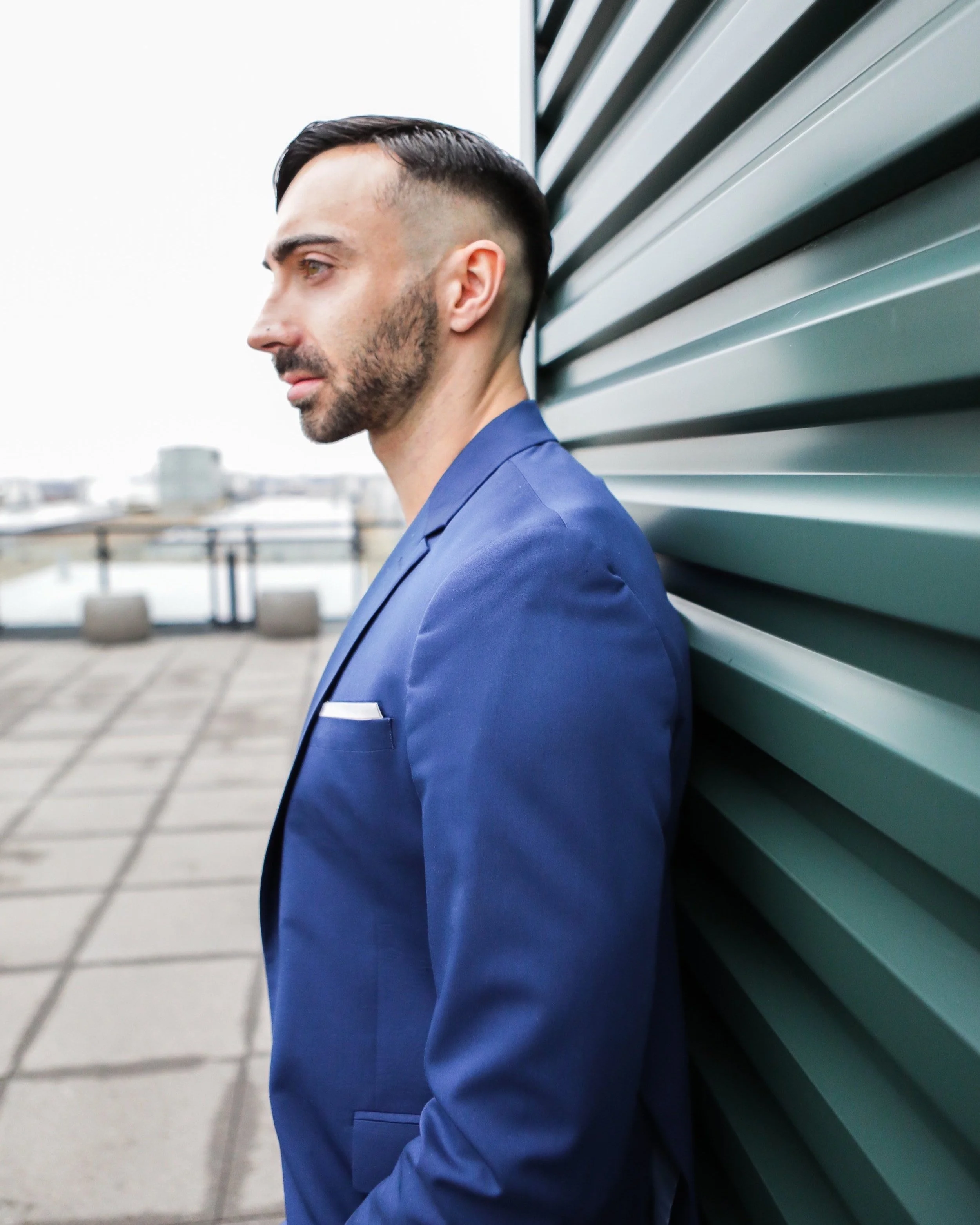 A man in a blue suit standing outdoors, leaning against a teal metal wall, with a blurred urban background.
