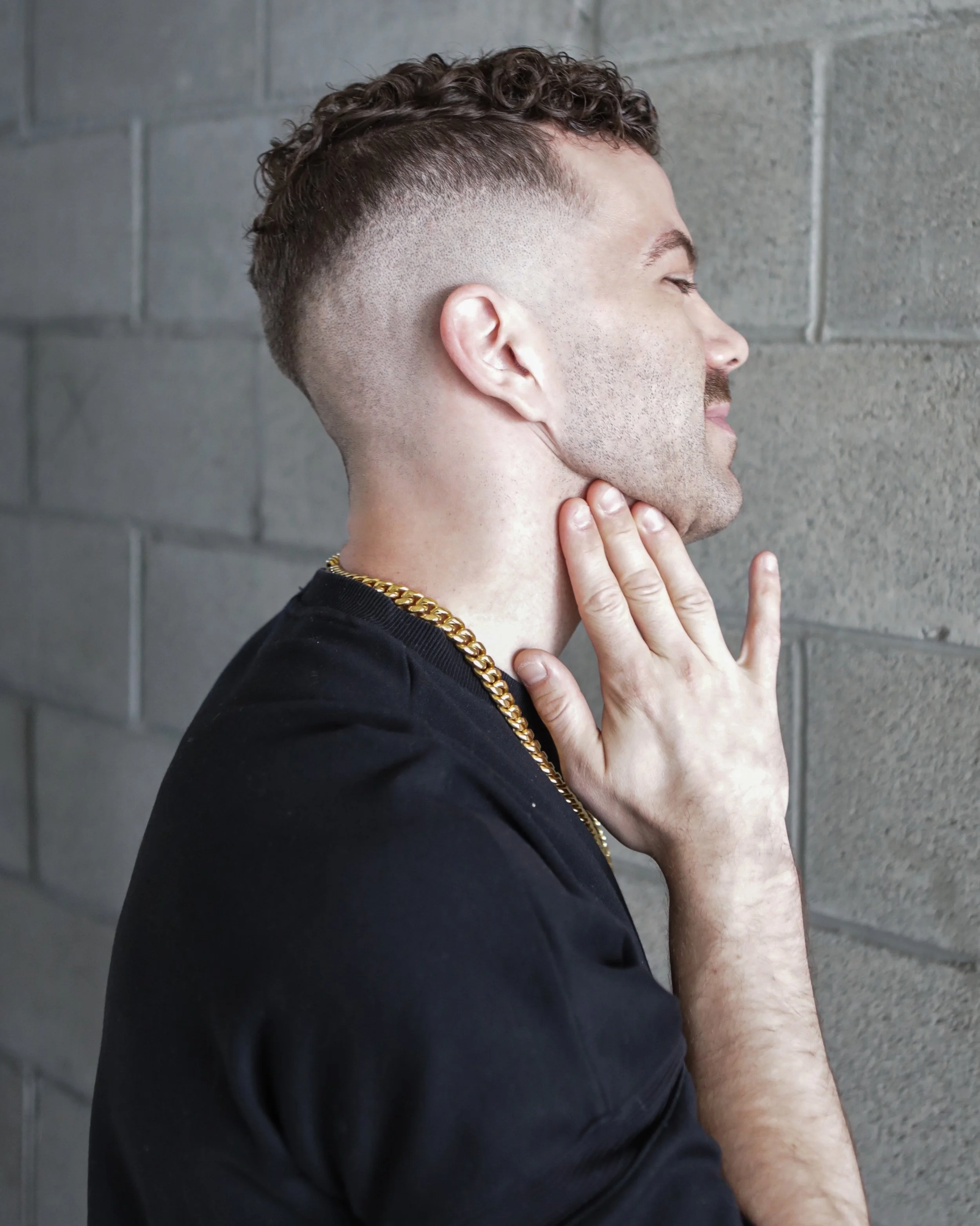 A young man with a curly hairstyle and buzzed sides, wearing a black shirt and a gold chain, is touching his neck and standing against a gray brick wall with eyes closed.