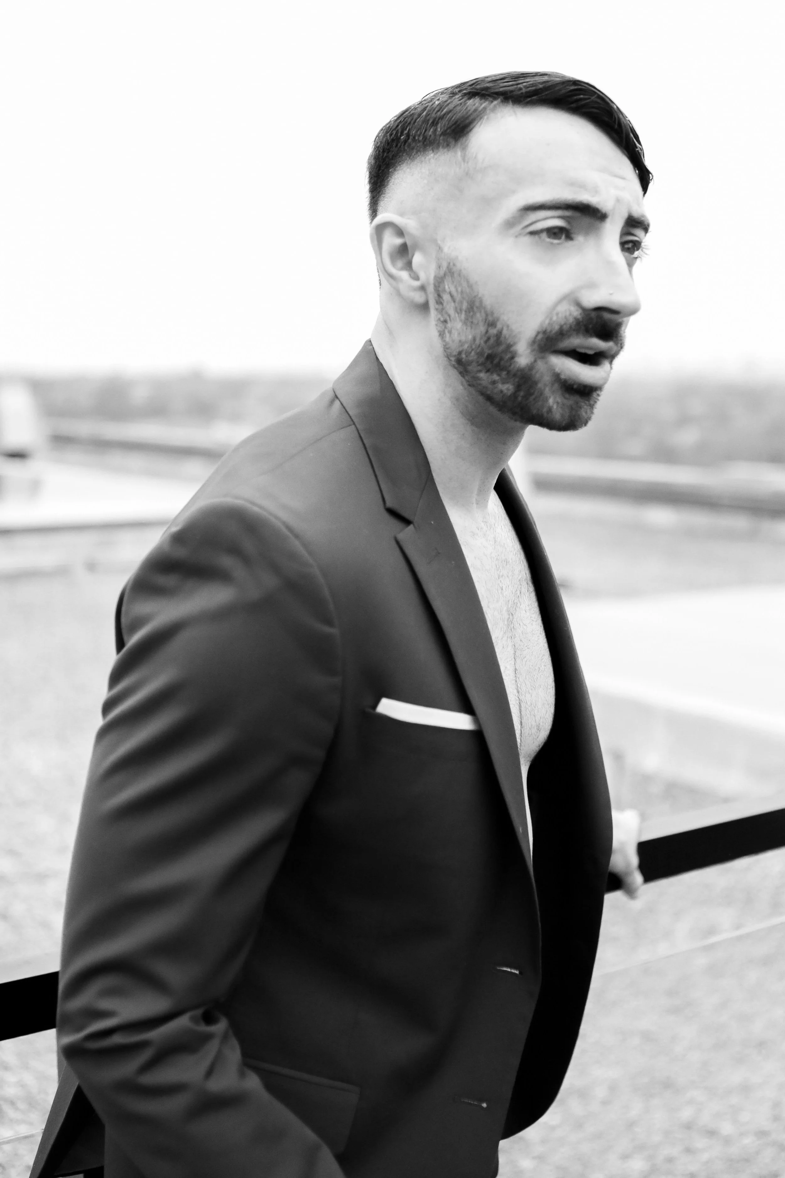 Black and white photo of a man with a beard and styled haircut, wearing a partially unbuttoned suit jacket with a pocket square, outdoors with a blurred background.