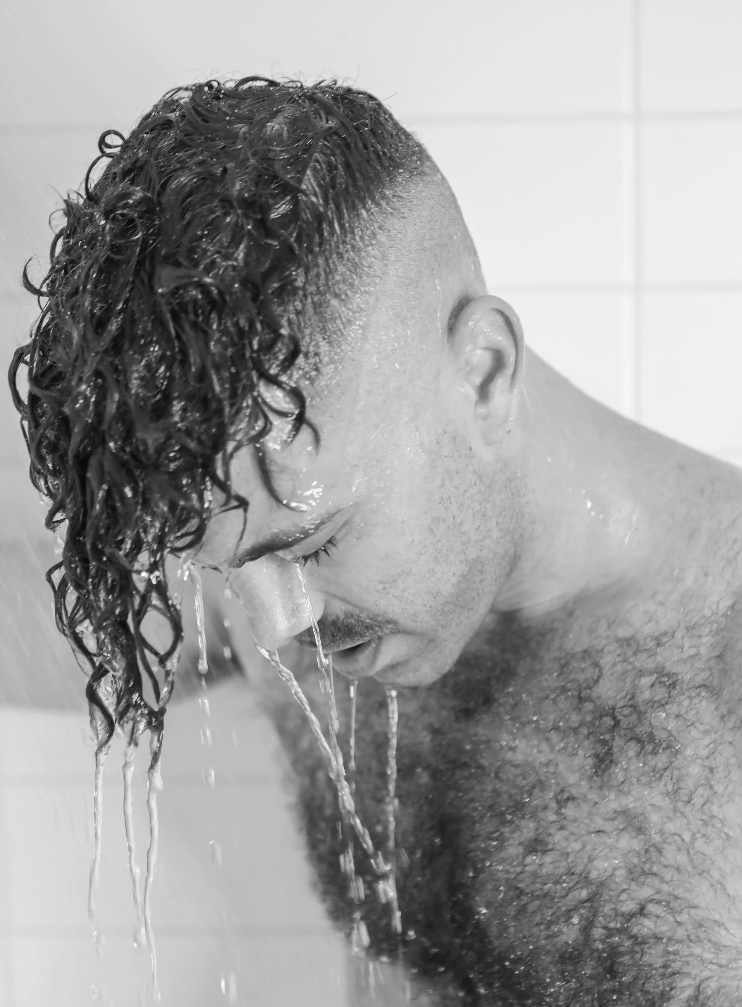 A man with wet, curly hair shaved on the sides, rinsing his face with water in a shower.