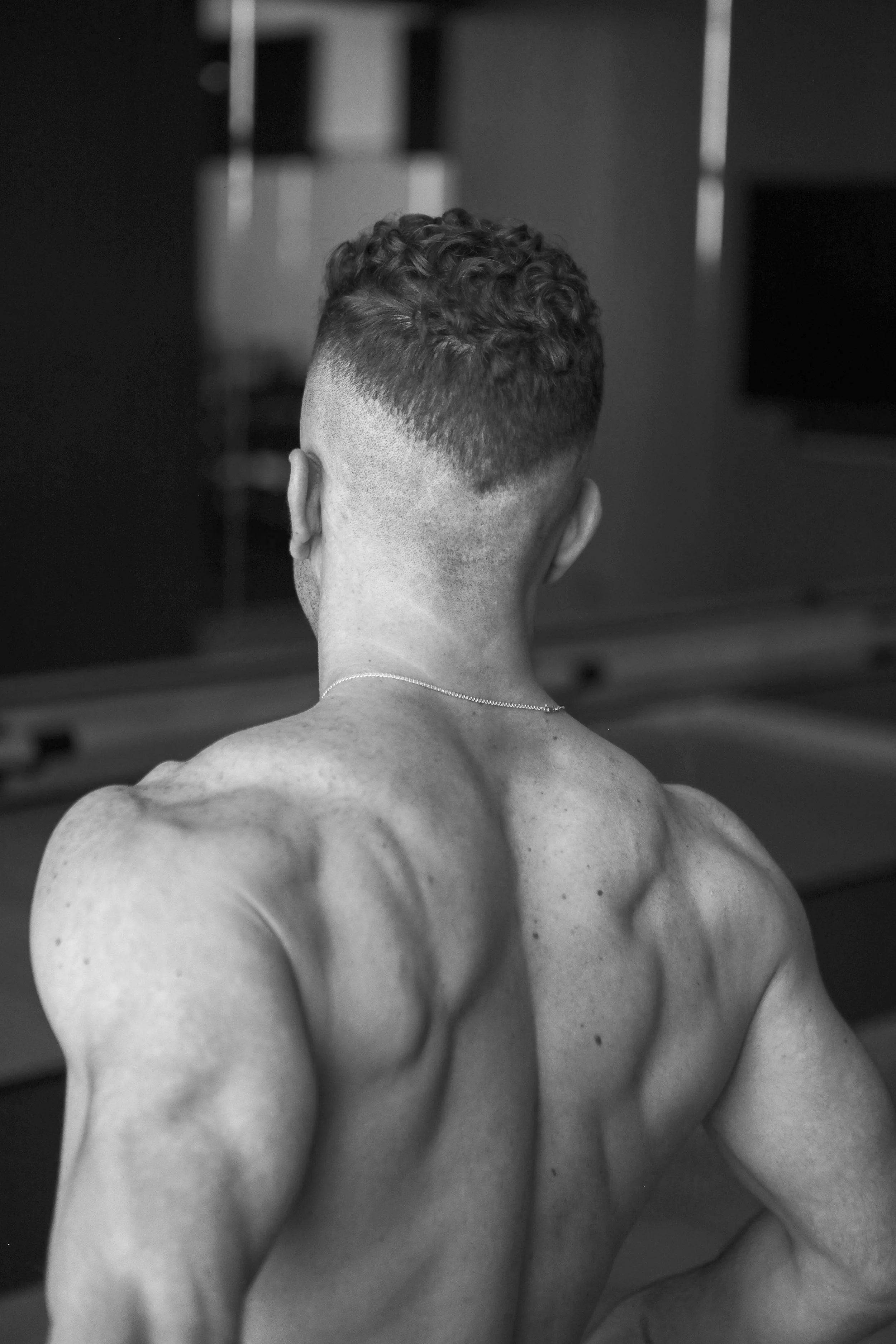 Back view of a muscular man with short curly hair, shirtless, in a black and white photo.