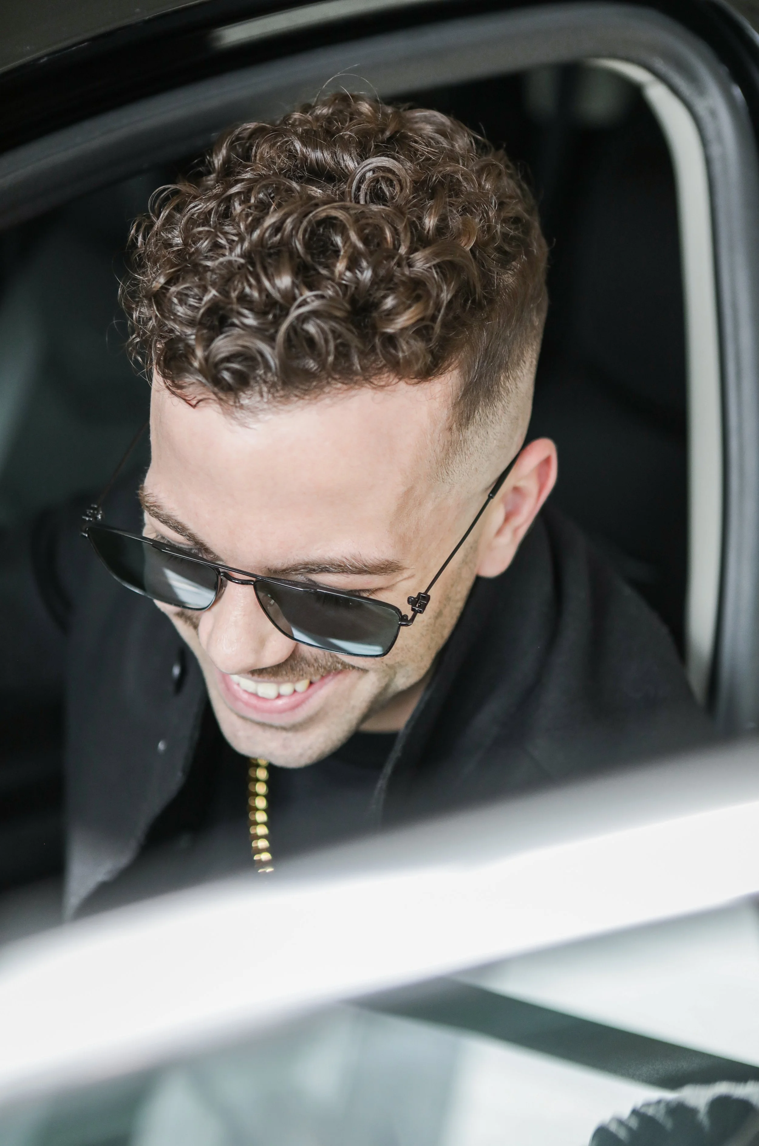 Smiling young man with curly hair wearing sunglasses, sitting in a car.