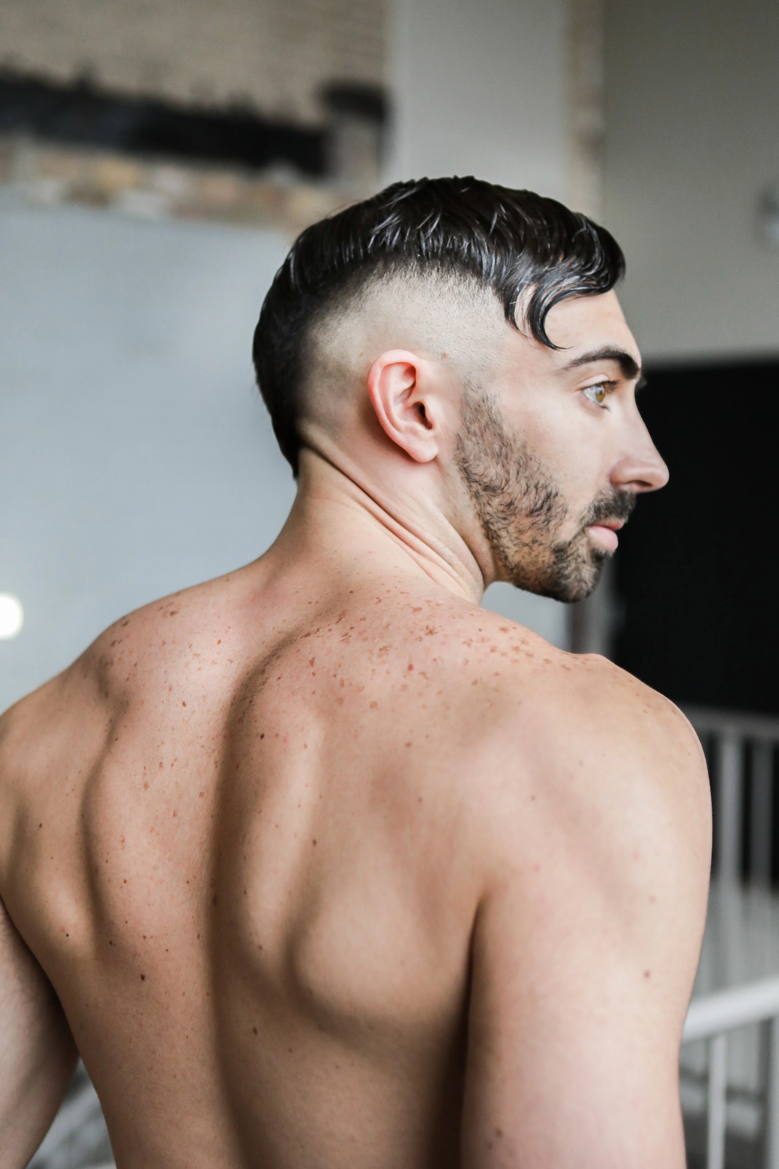 A man with dark, slicked-back hair and a beard showing a close-up of his bare back and shoulder, highlighting freckles and skin texture.